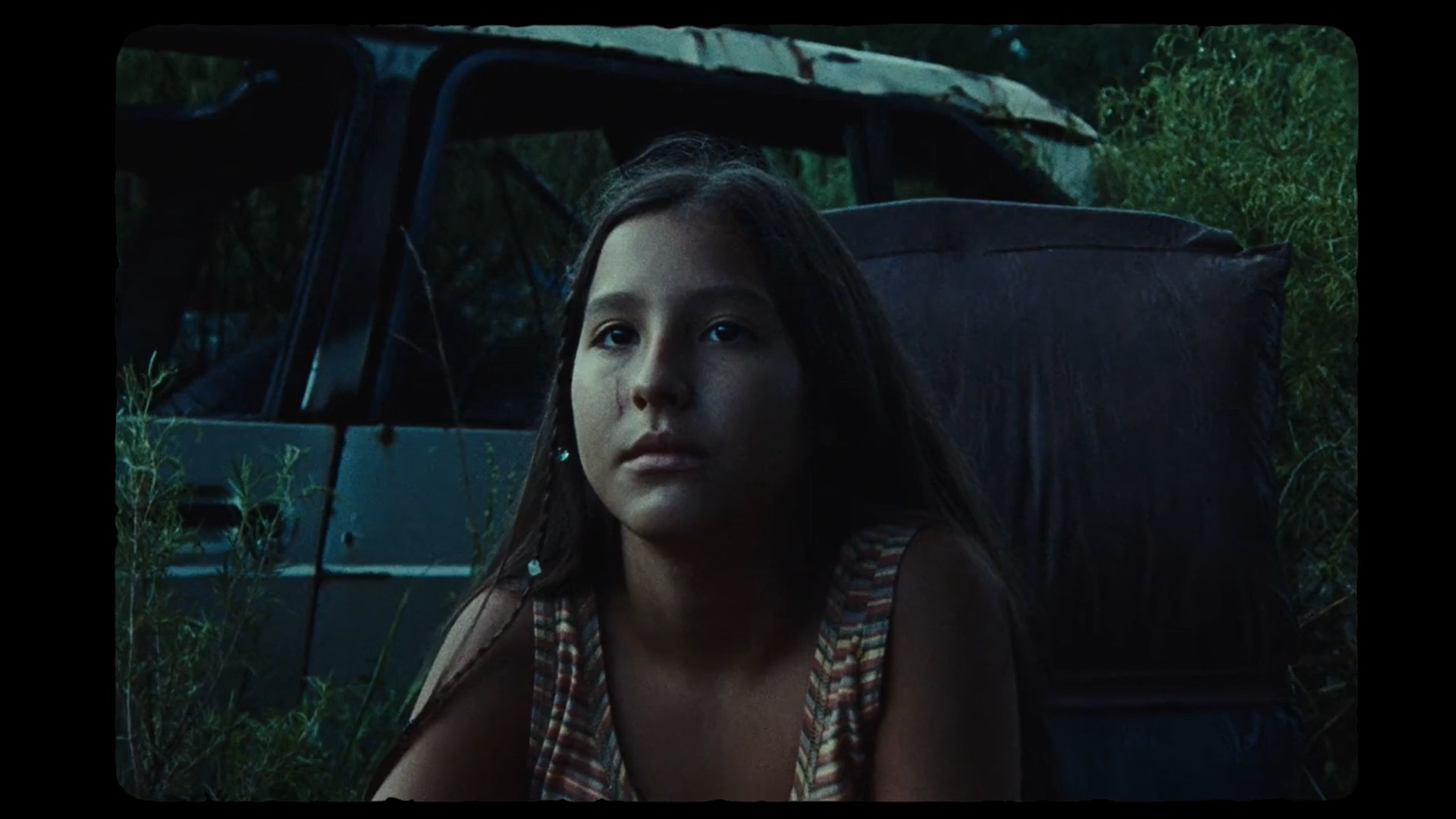 A young woman with long dark hair and a striped shirt kneels in front of a damaged vehicle in a grassy outdoor area during dusk or nighttime.