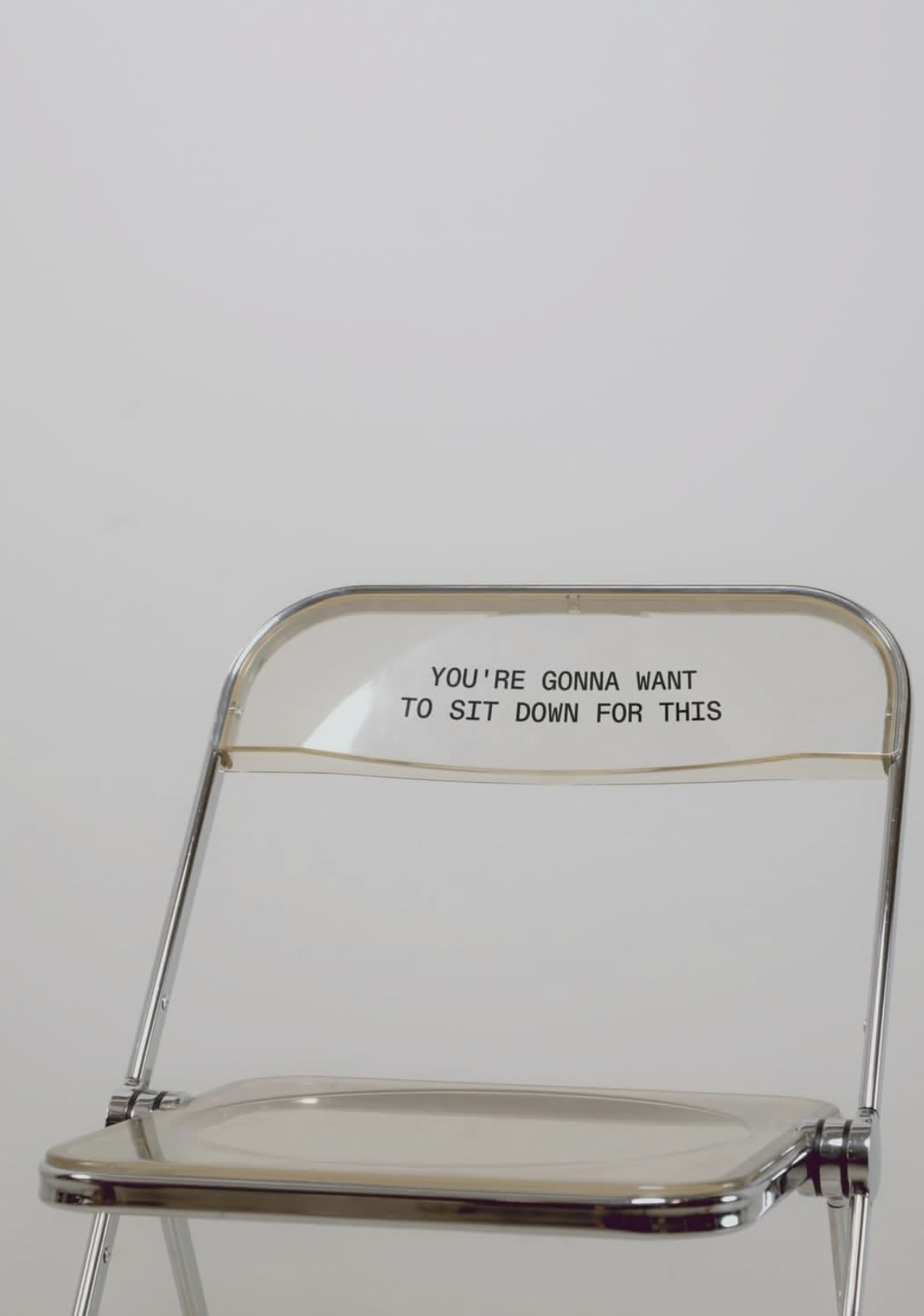 A transparent plastic chair with a sign on the backrest that reads, 'YOU'RE GONNA WANT TO SIT DOWN FOR THIS' against a plain white background.