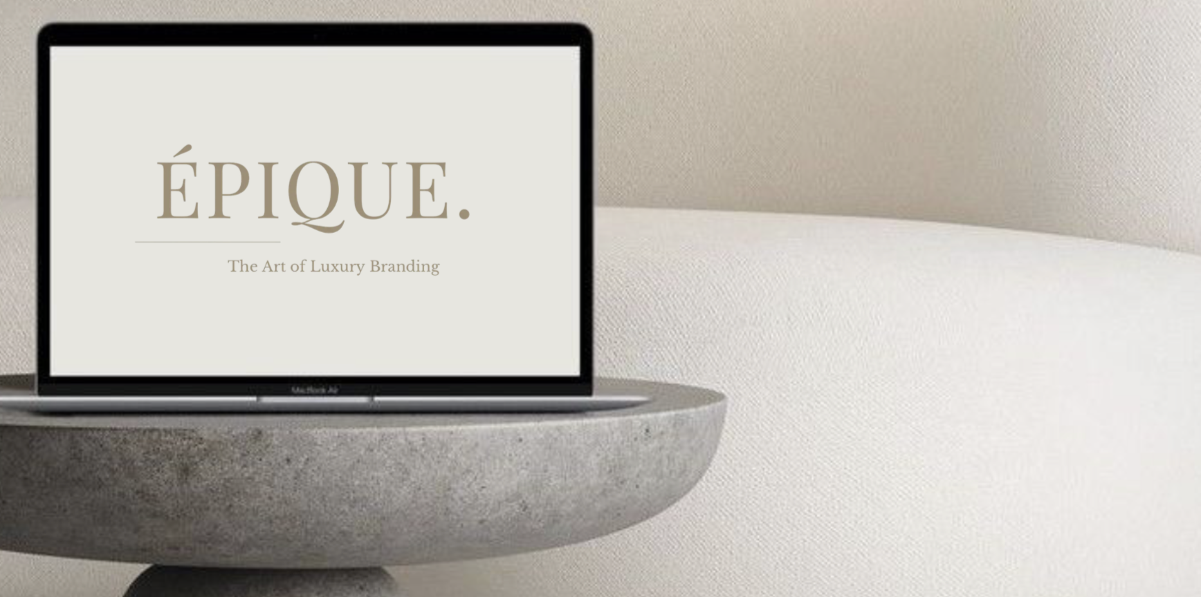 A MacBook Air laptop displaying the words 'ÉPIQUE. The Art of Luxury Branding' on a minimalist beige background, placed on a curved, textured gray concrete tabletop against a light beige wall.