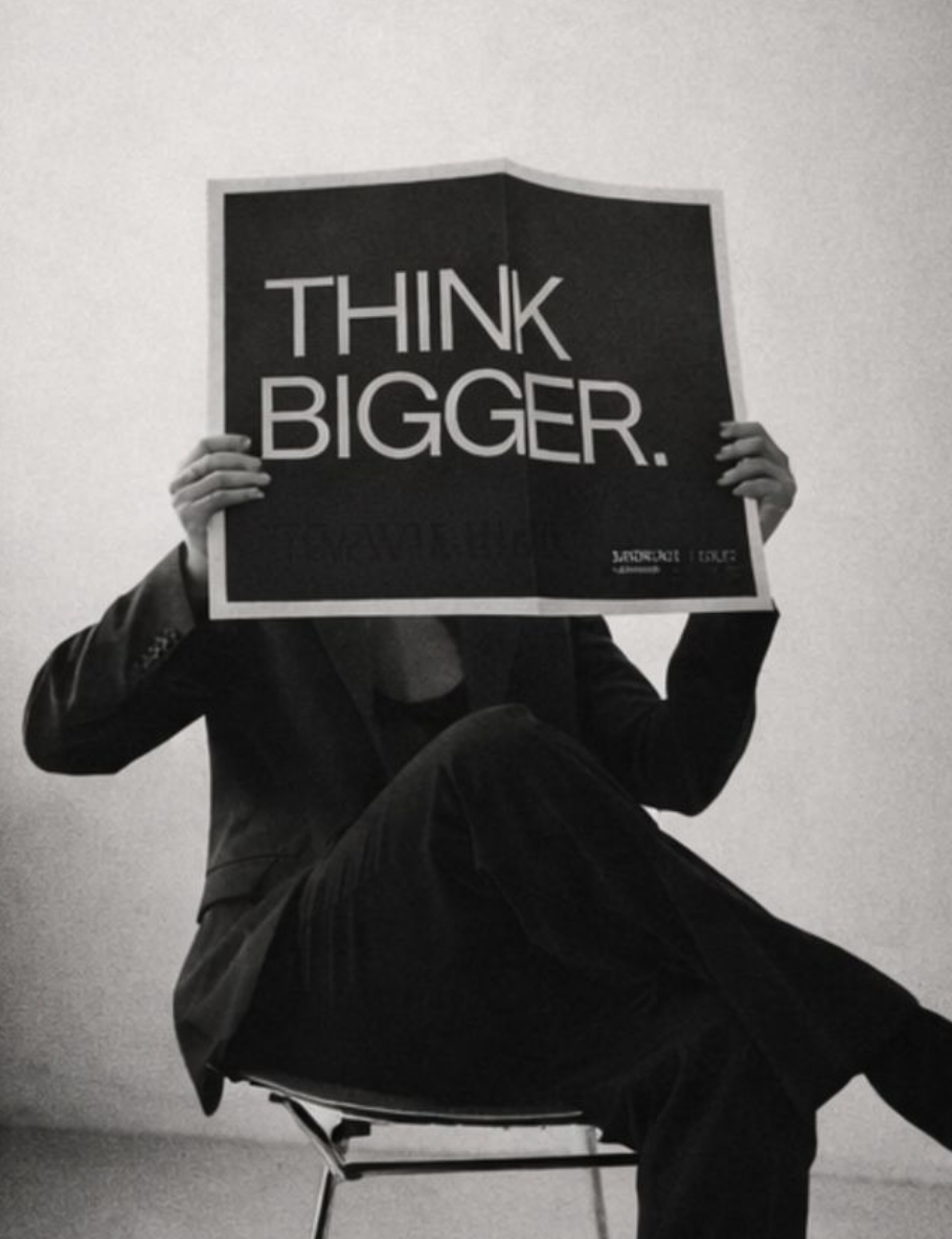 Person sitting cross-legged, holding a sign that says 'THINK BIGGER' in front of their face, in a black and white photo.