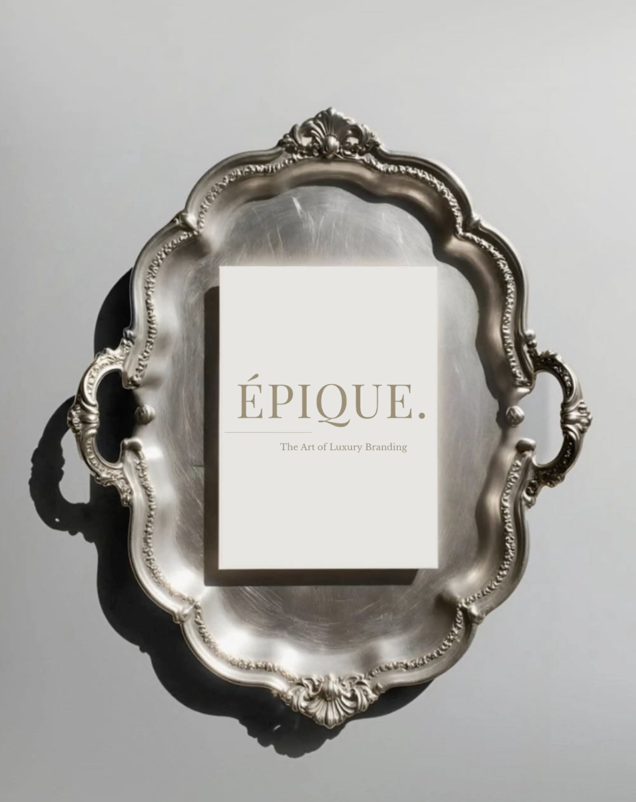An ornate silver mirror with intricate detailing. A white book or catalog titled 'ÉPIQUE. The Art of Luxury Branding' is placed on the mirror. The background is a plain, light-colored wall.