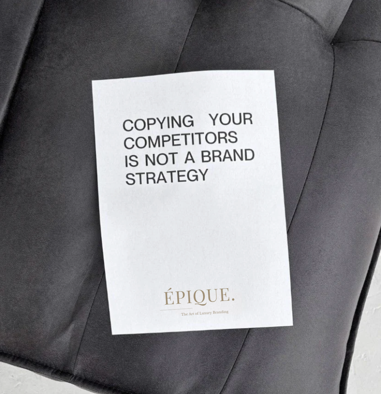 A white paper with the text "COPYING YOUR COMPETITORS IS NOT A BRAND STRATEGY" in black, placed on a black leather surface. The bottom of the paper features the logo and tagline of Épique, a luxury branding company.