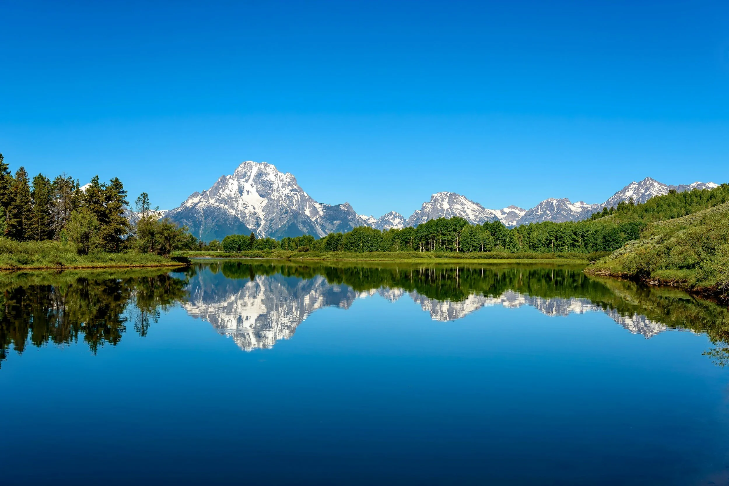 Snow-capped mountains reflected in a calm lake, with green trees along the shoreline under a clear blue sky.
