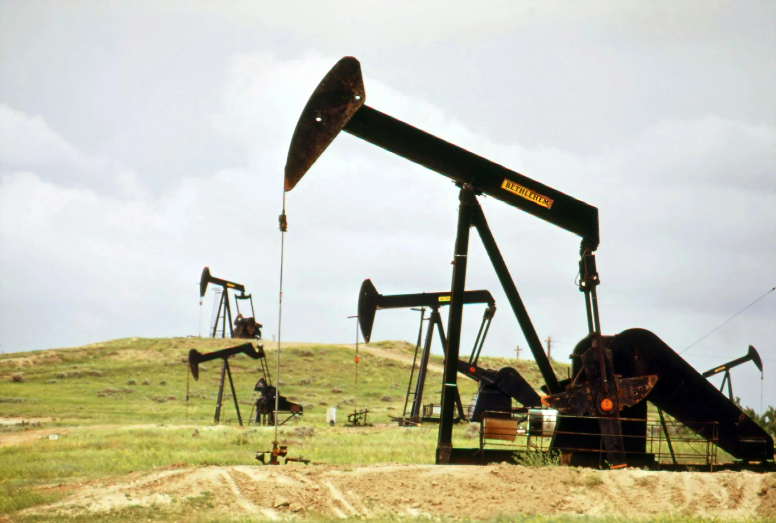 Multiple oil pump jacks on a grassy landscape under a cloudy sky, with a hill in the background.