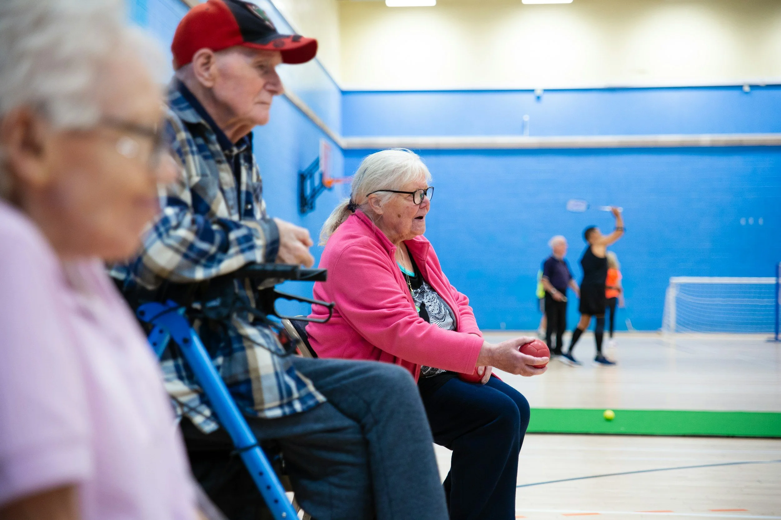 An older woman wearing glasses and a pink jacket is sitting and holding a red ball in her hands. She is in a gymnasium with a blue wall in the background, where children are playing with a tennis ball.