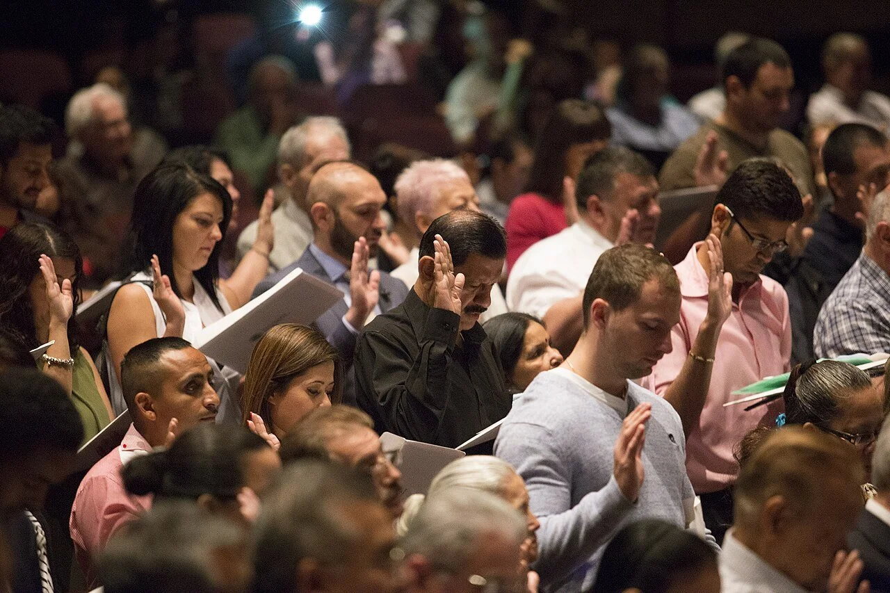 A group of diverse people standing outdoors with their right hands raised, participating in a naturalization ceremony.