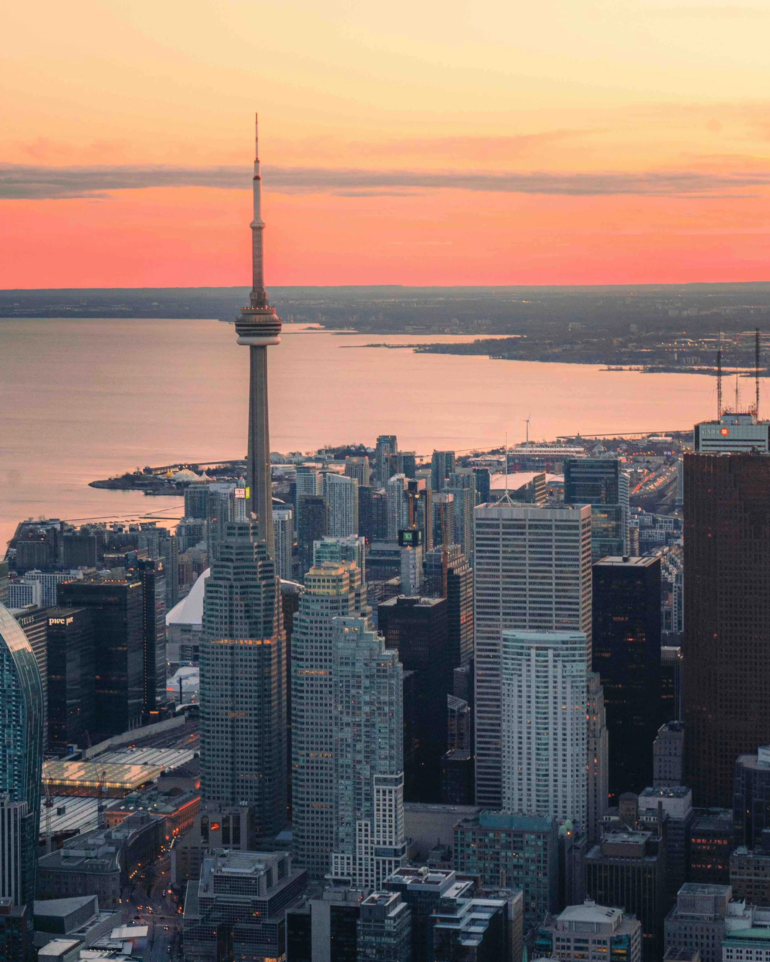 City skyline at sunset featuring the CN Tower rising above downtown skyscrapers with Lake Ontario visible in the background.