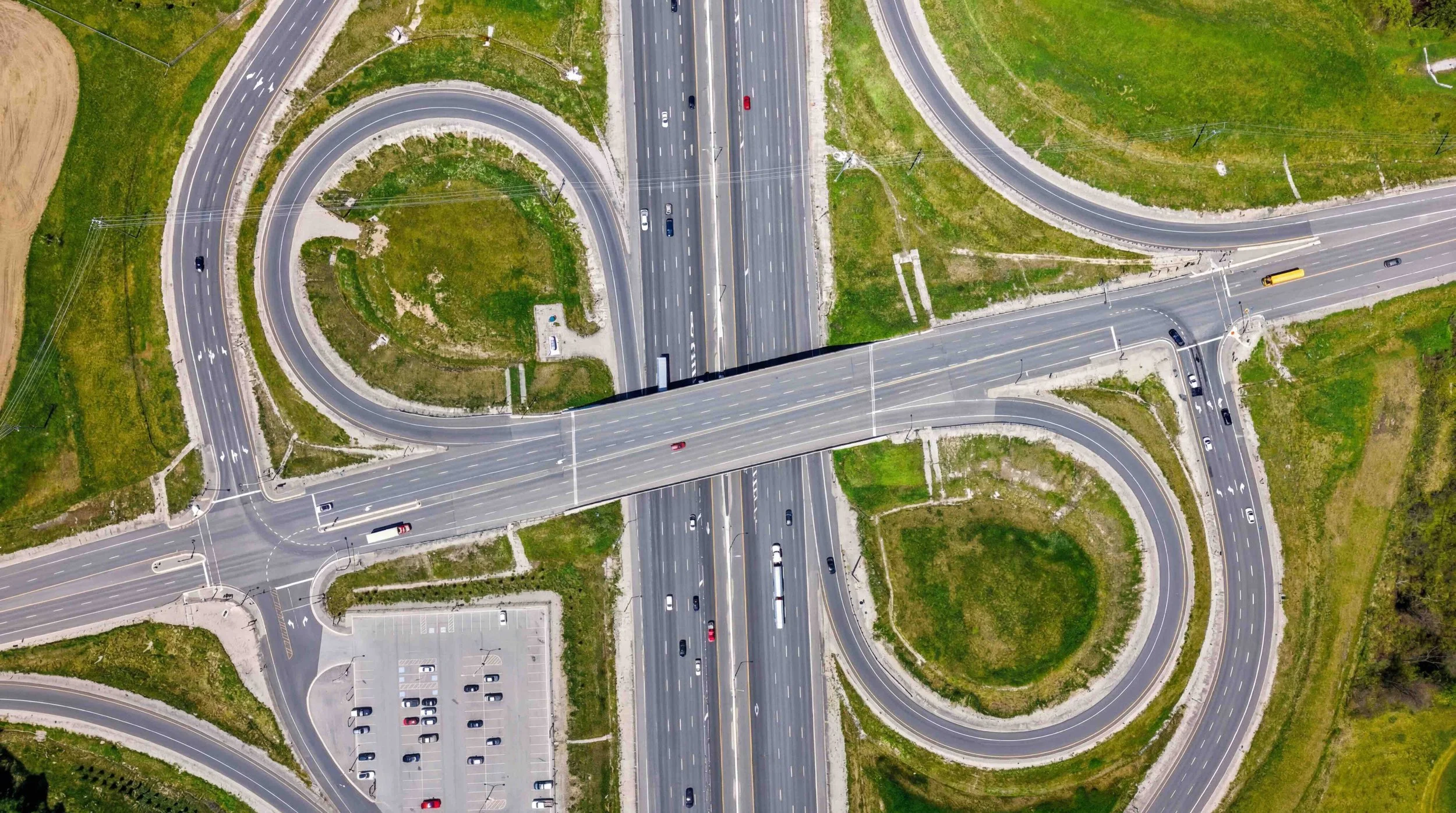 Aerial view of a large highway interchange with looping ramps and an overpass crossing a multi-lane freeway, surrounded by green grass and light traffic.