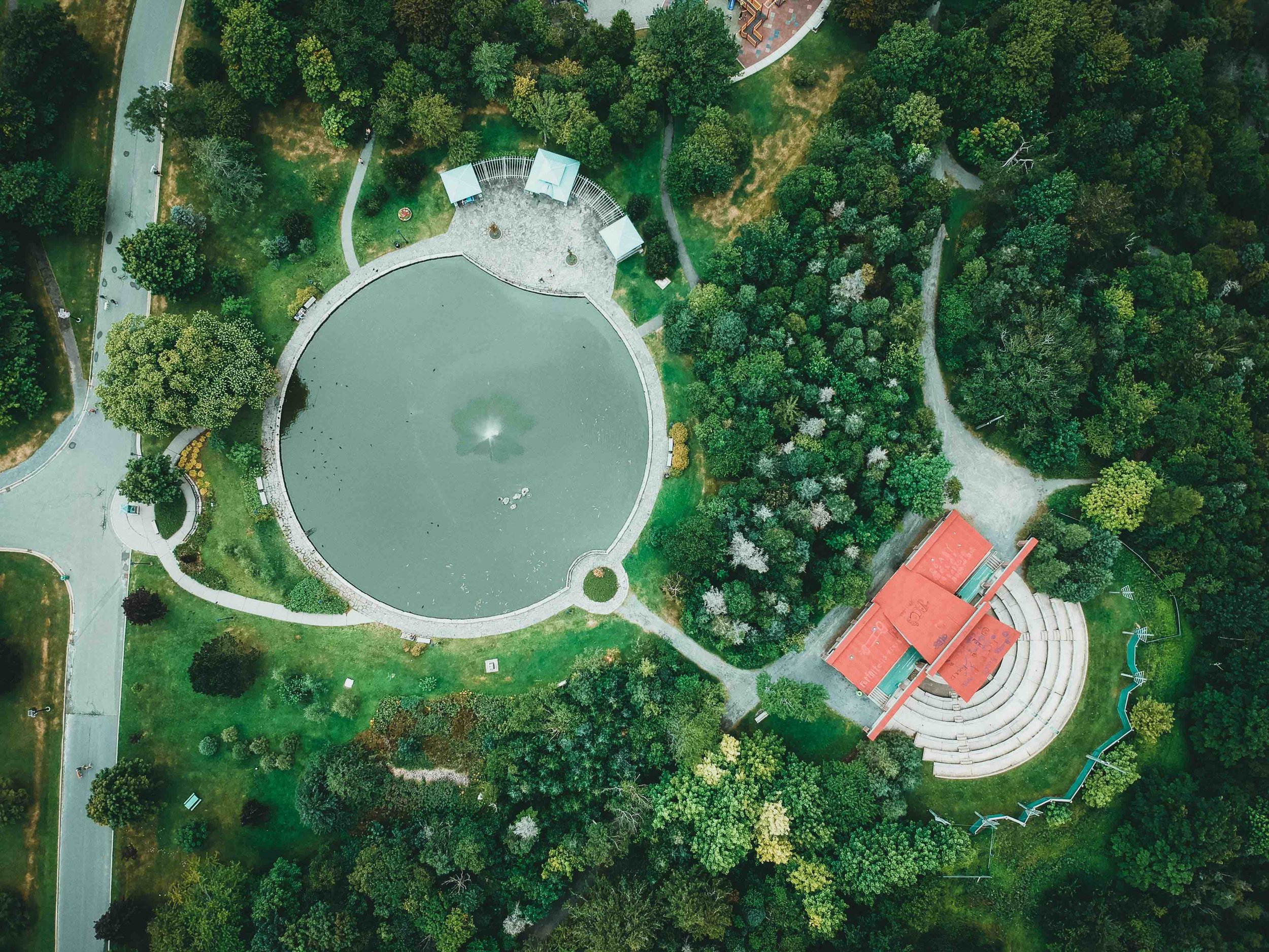 Aerial view of a circular pond surrounded by walking paths, dense trees, and a small amphitheater-like structure in a landscaped park.