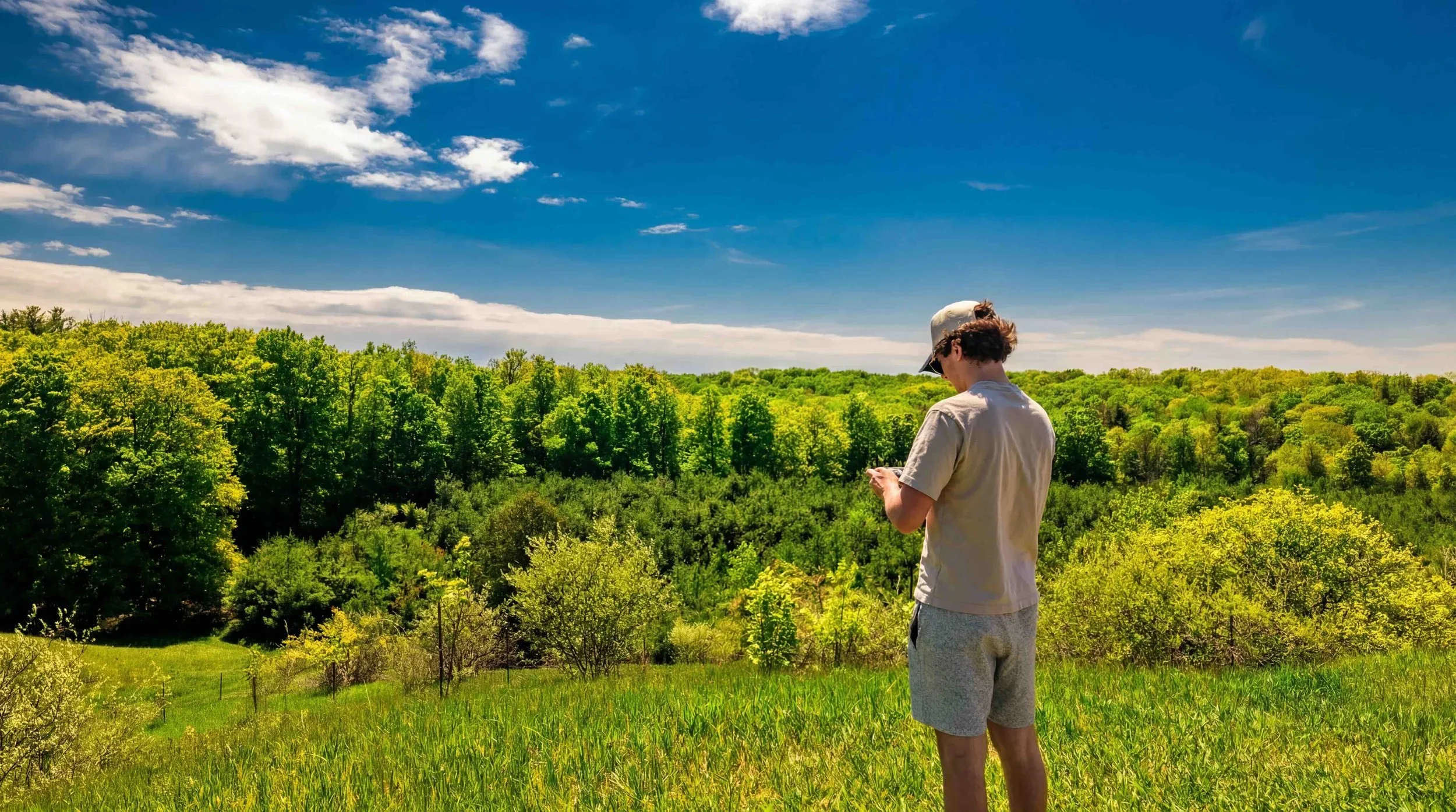 Marcus Gingl standing in a grassy field, looking down at a phone while overlooking a lush green forest beneath a bright blue sky with scattered clouds.