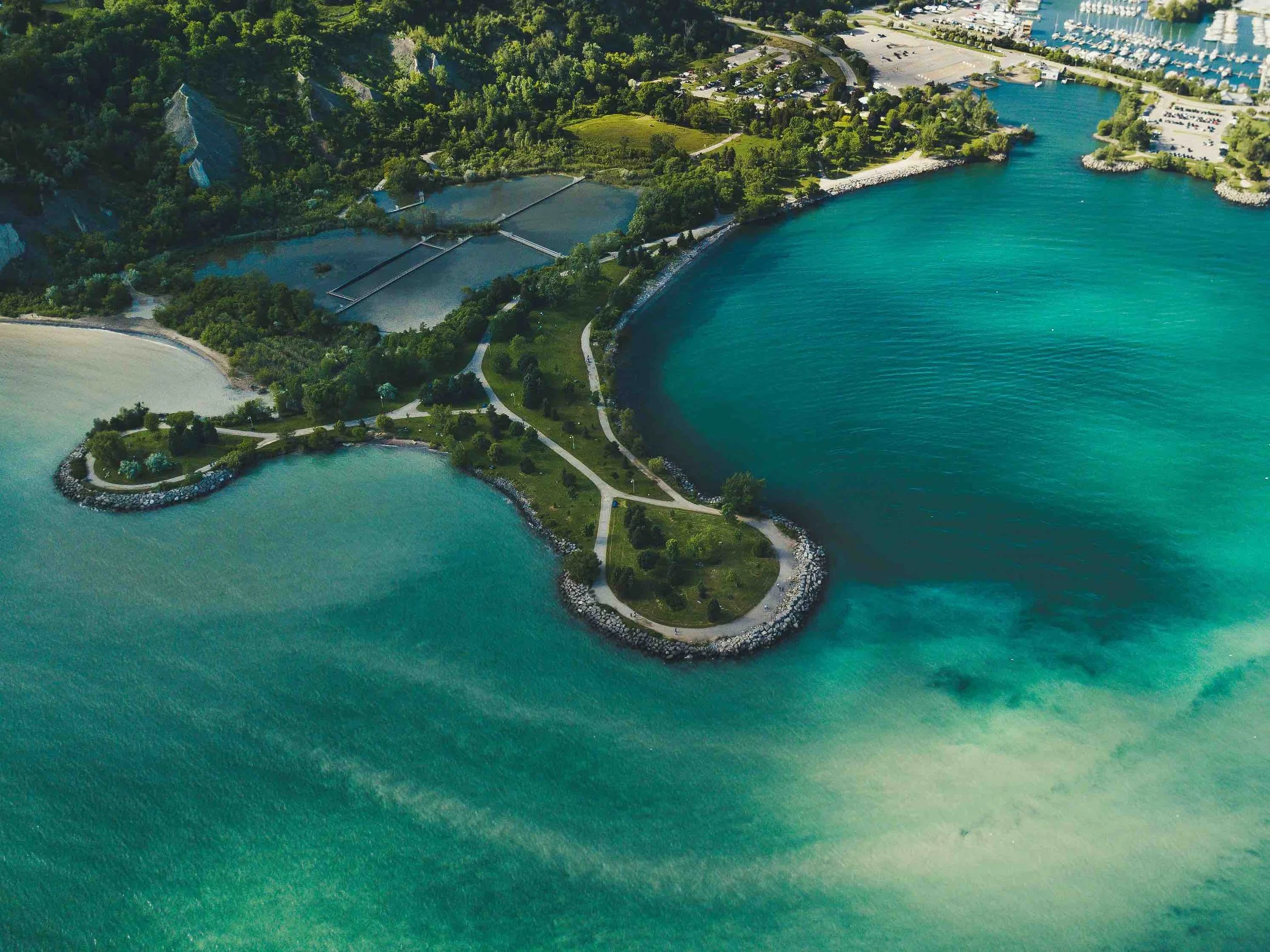 Drone view of a turquoise bay with a small tree-covered island connected to a curving shoreline, bordered by clear blue-green water.