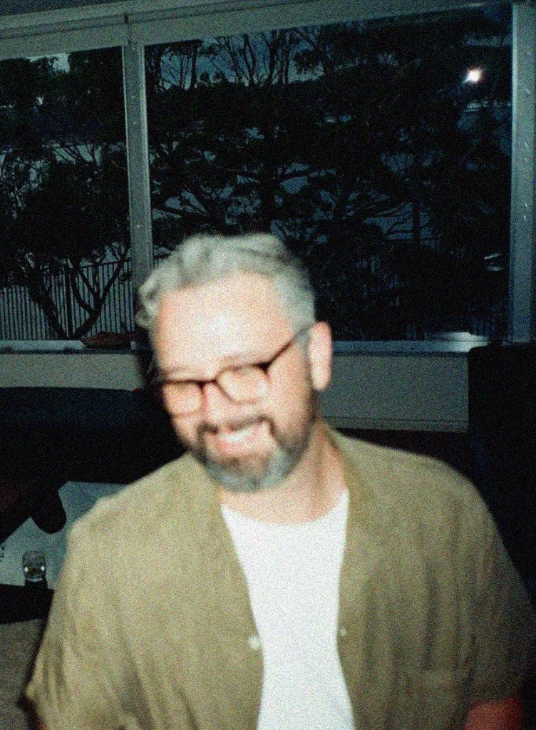 A smiling man with gray hair, beard, and glasses, wearing a brown jacket and white shirt, sitting indoors in front of a window showing trees and a dark sky. Graphic designer, artist and creative.