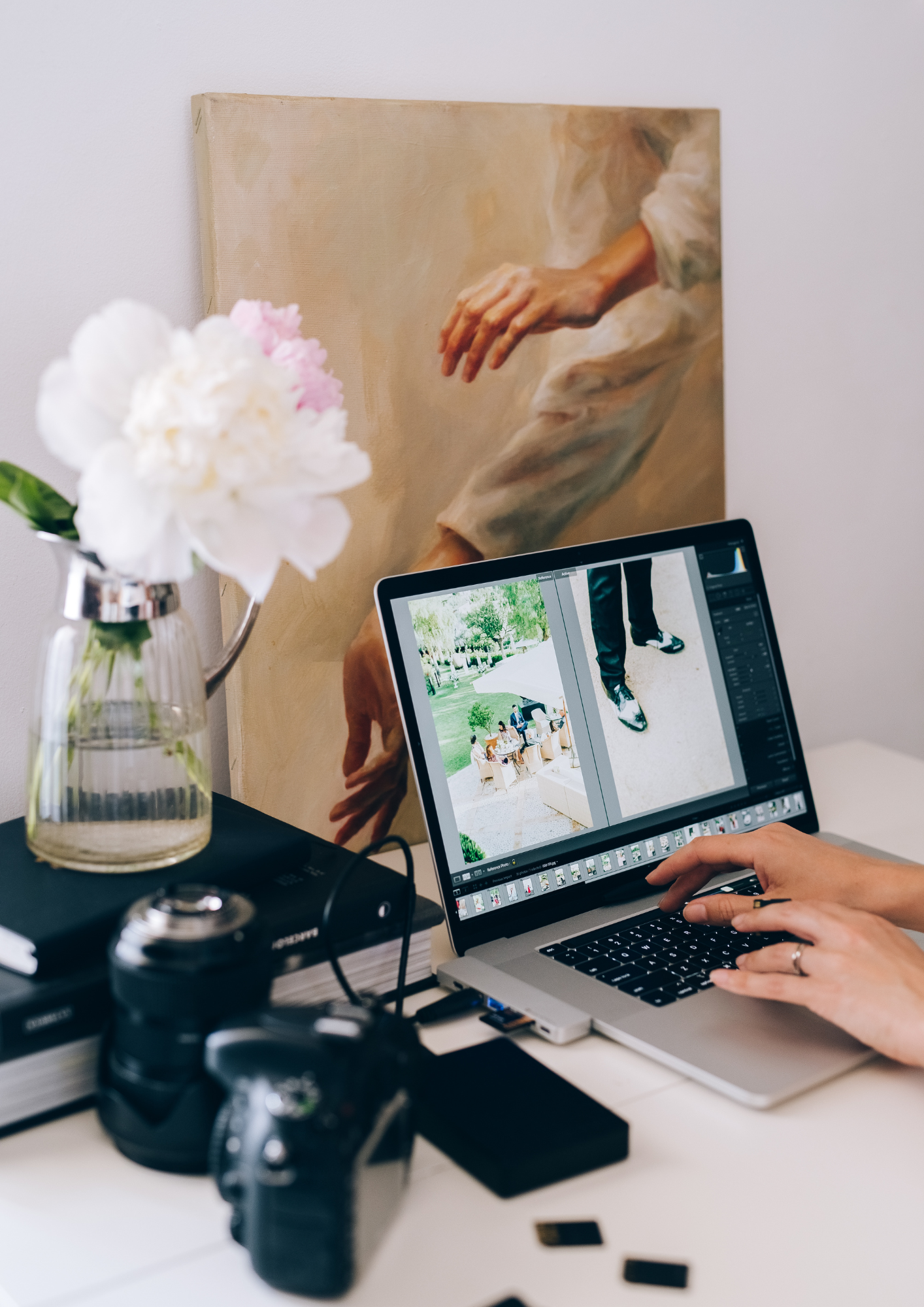 A person working on photo editing on a laptop at a desk, with a painting of a hand in the background and a flower vase on the side.