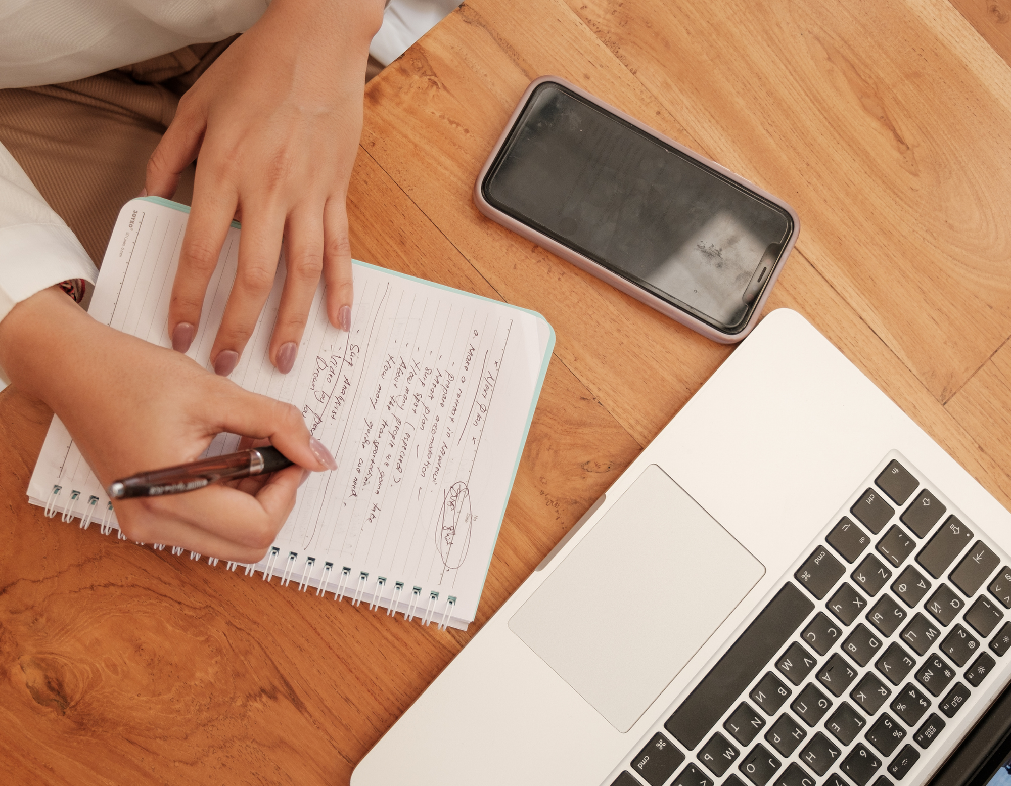 A person writing in a notebook with handwritten notes, sitting at a wooden desk with a smartphone, a laptop, and a piece of paper nearby.