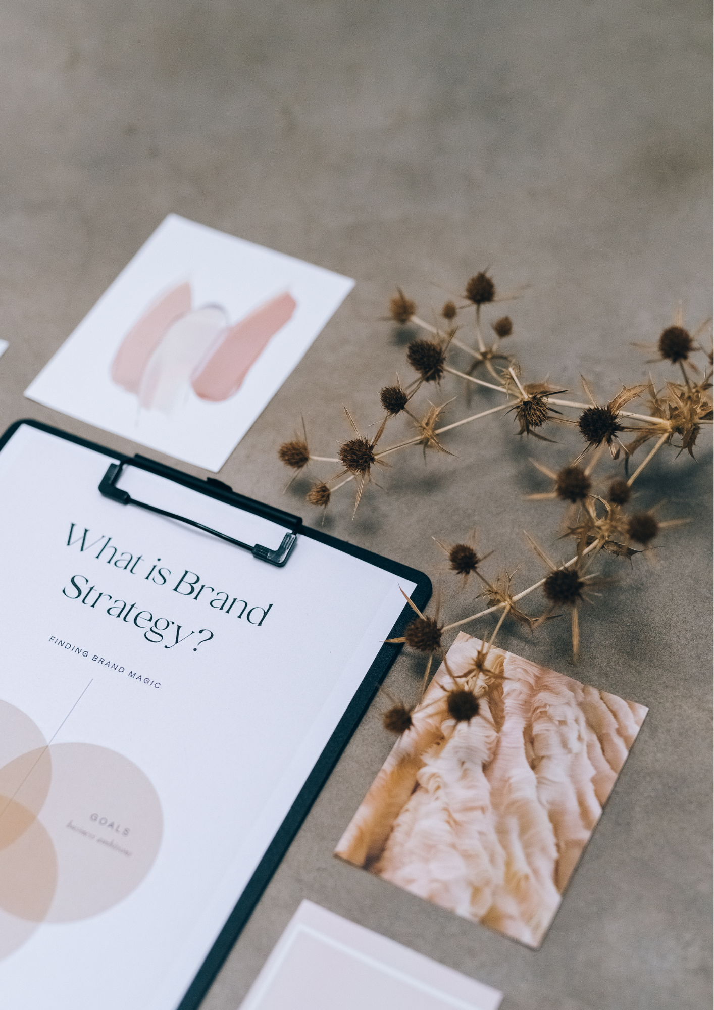 A flat lay of a study or planning workspace showing a paper titled 'What is Brand Strategy?' with a color palette and goals diagram, a painted photo of a lion, a dried plant, and other print materials on a grey surface.