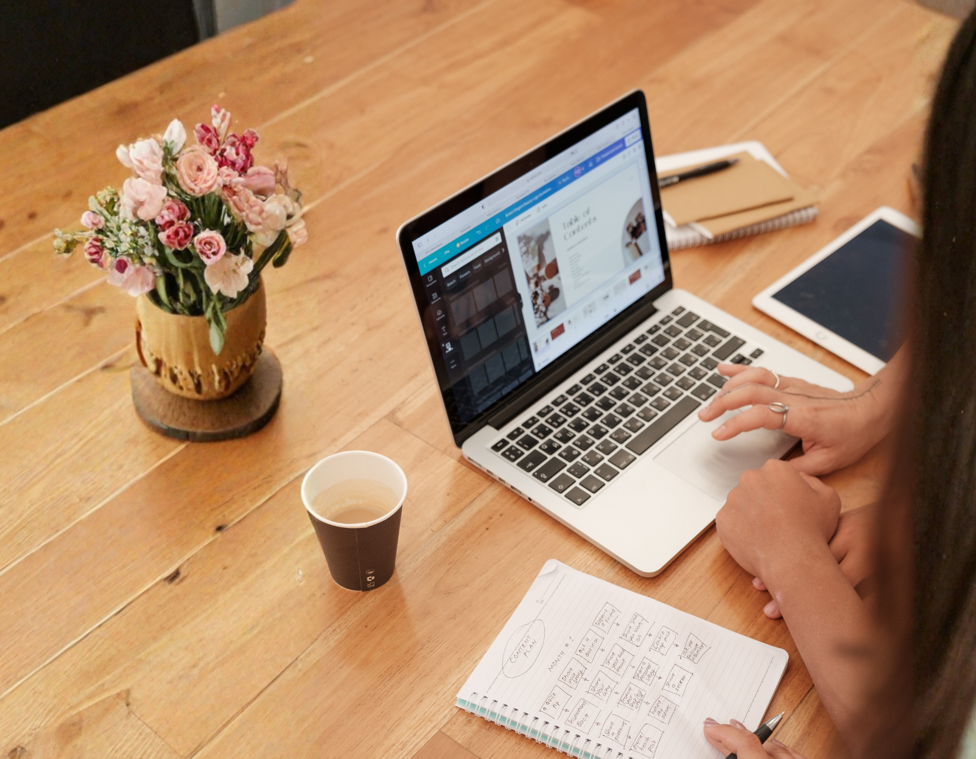 A person working on a laptop at a wooden table with a notebook, pen, and a cup of coffee. There is a vase with pink and white flowers on the table.