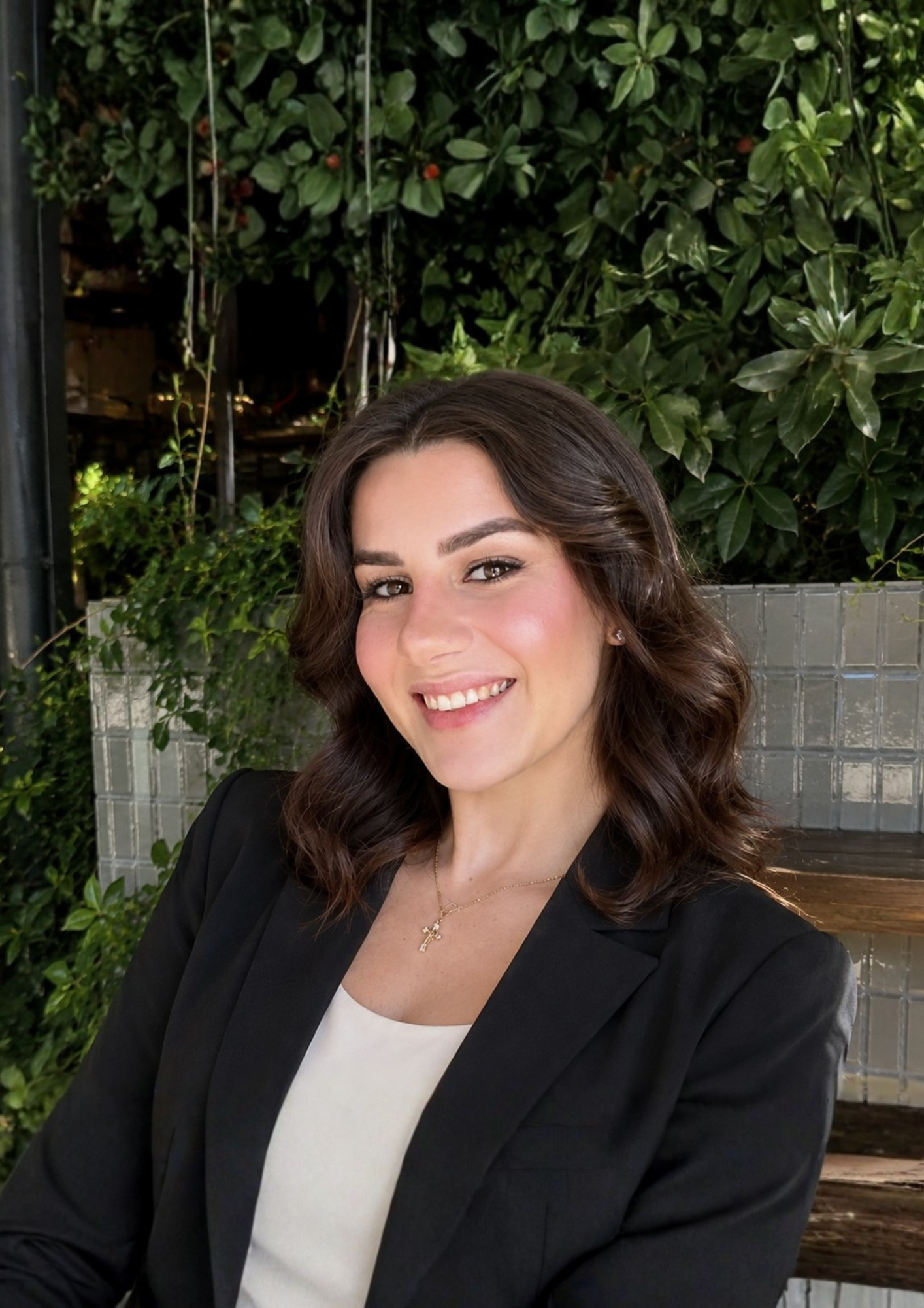 A woman with brown wavy hair smiling, wearing a black blazer, white top, a gold cross necklace, sitting outdoors with green foliage behind her.