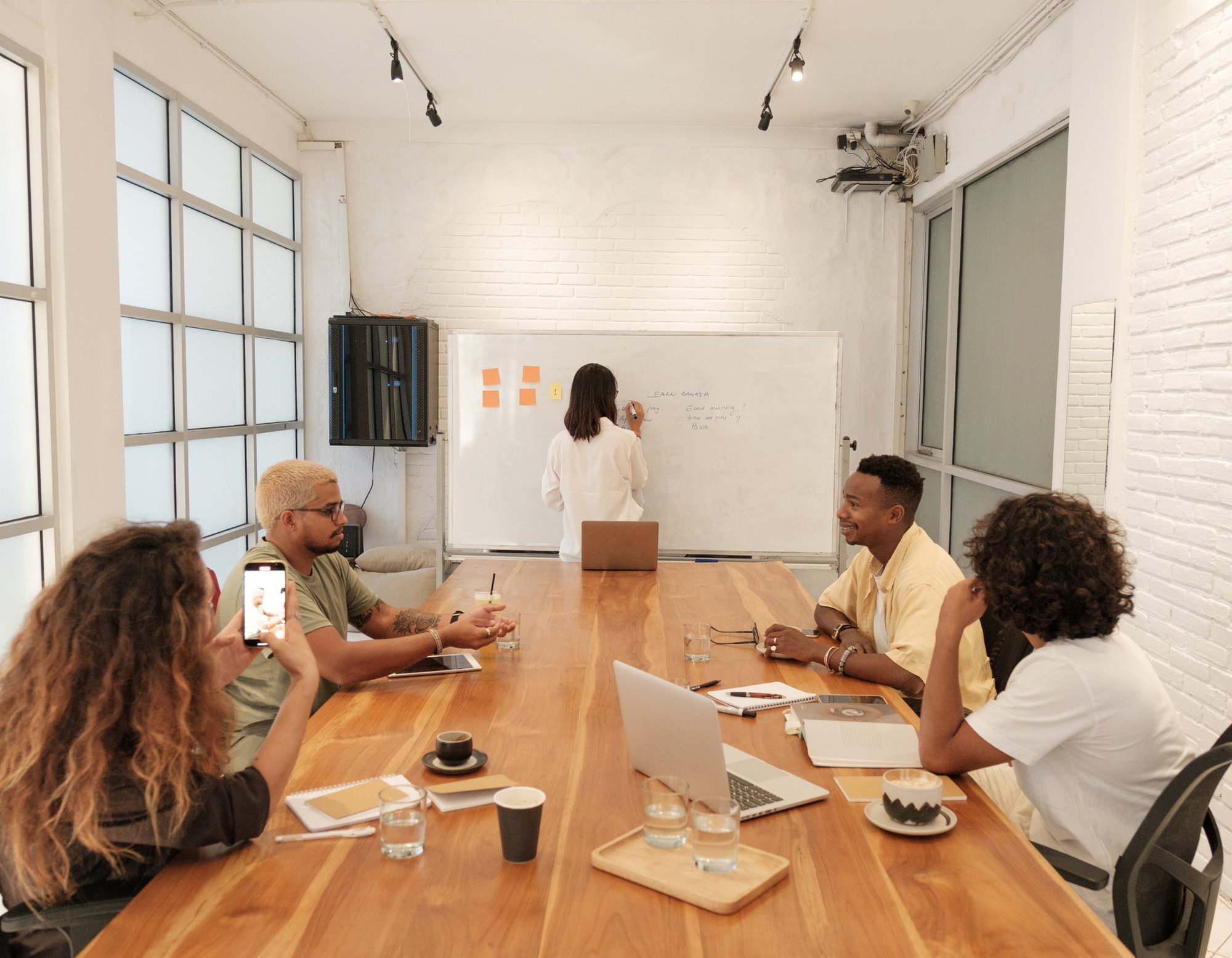 Group of people in a meeting room, seated around a wooden table with laptops, notebooks, and drinks, listening to a woman at the whiteboard.