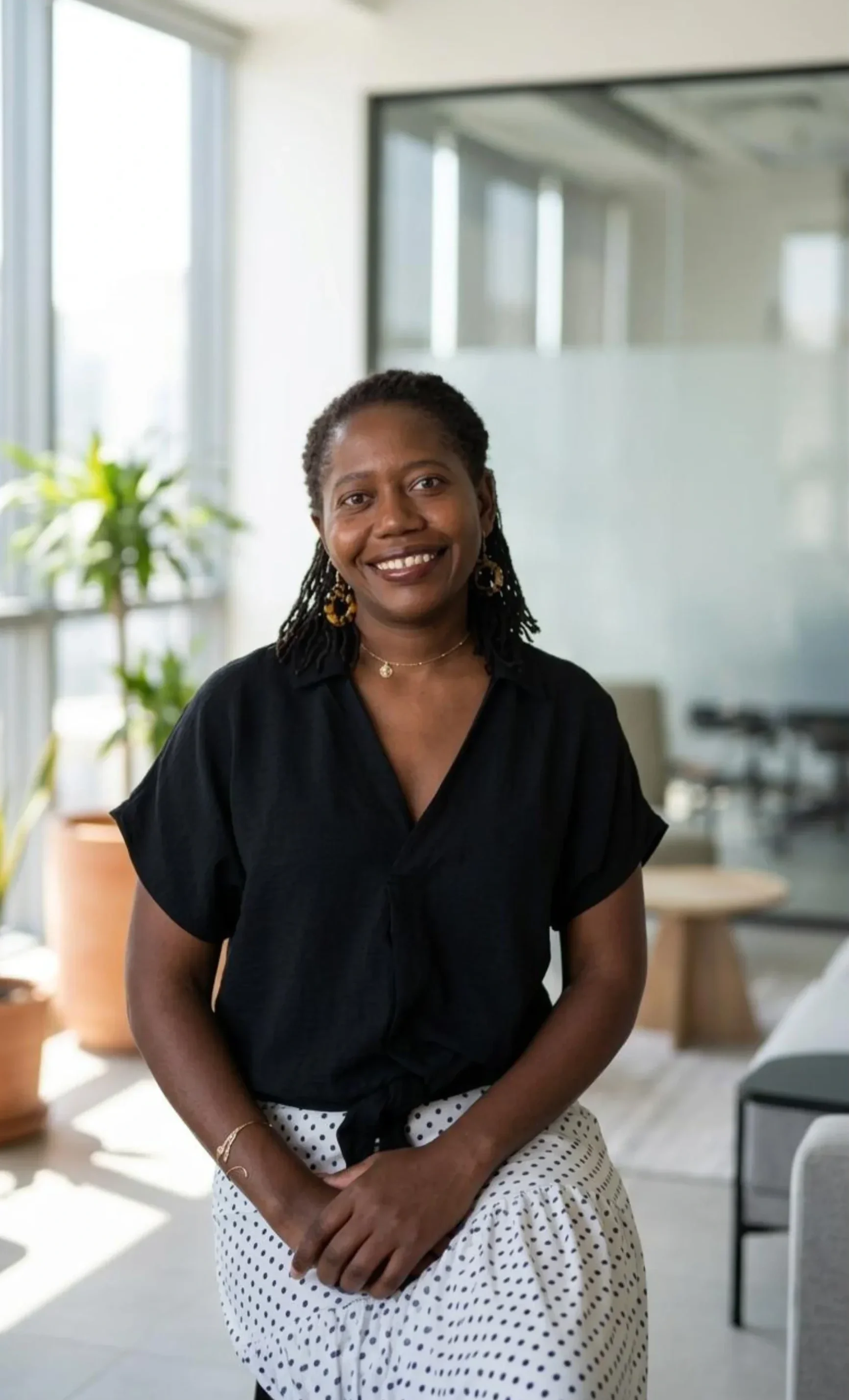 Smiling black woman with short curly hair wearing a black blouse and polka dot skirt, standing in a bright modern office space with large windows and indoor plants.