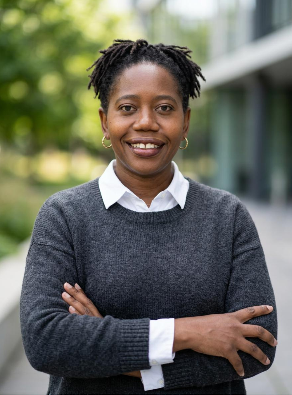 A confident woman with short dreadlocks and hoop earrings smiling outdoors with crossed arms, wearing a dark gray sweater over a white shirt, in front of greenery and a modern building.