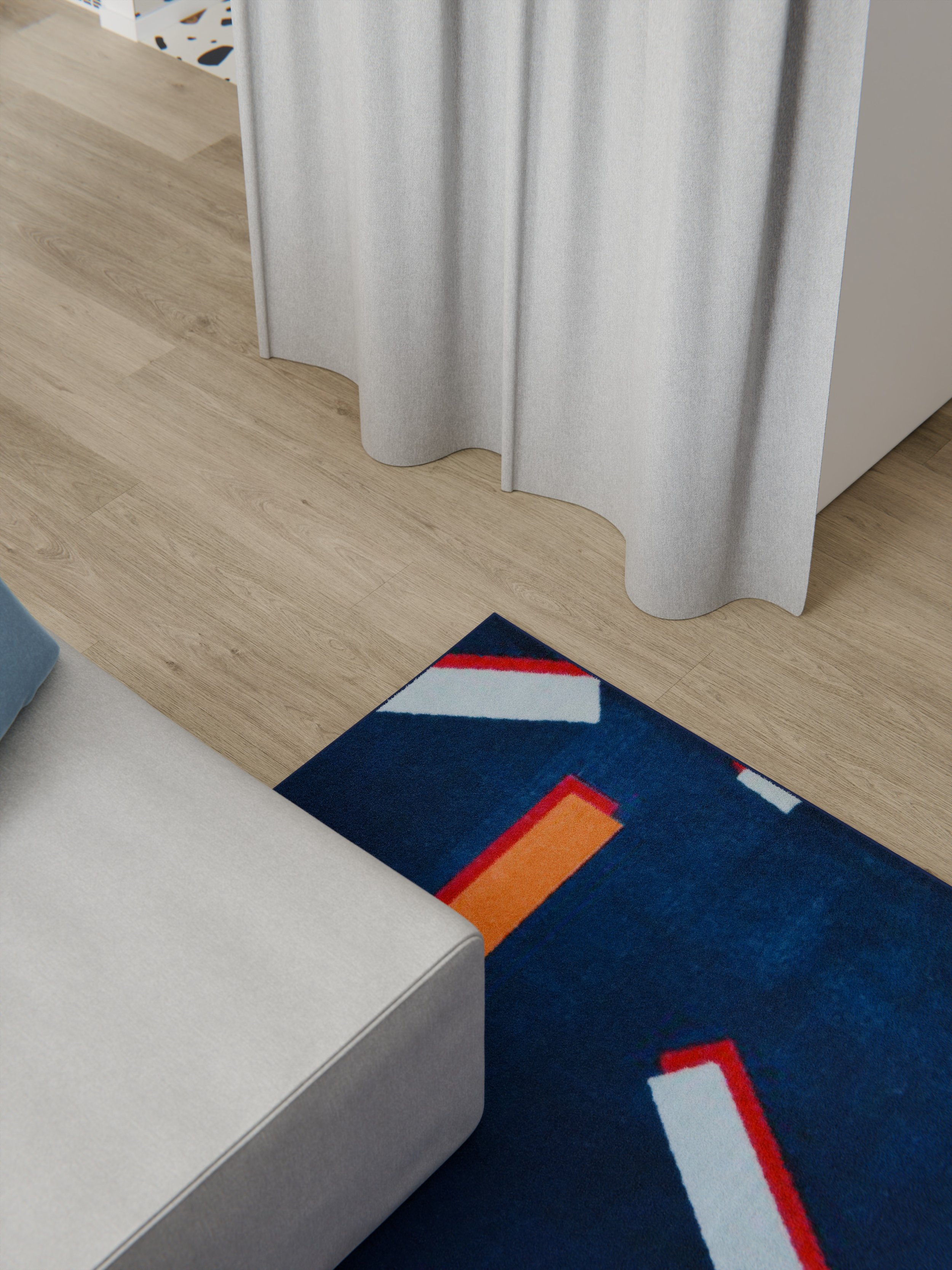 La foto mostra un angolo di un soggiorno con un tappeto colorato con motivi geometrici, un divano grigio chiaro e una tenda grigia. Il pavimento è in legno chiaro, e si intravede una zona con piastrelle bianche nere e blu nella parte superiore dell'immagine.