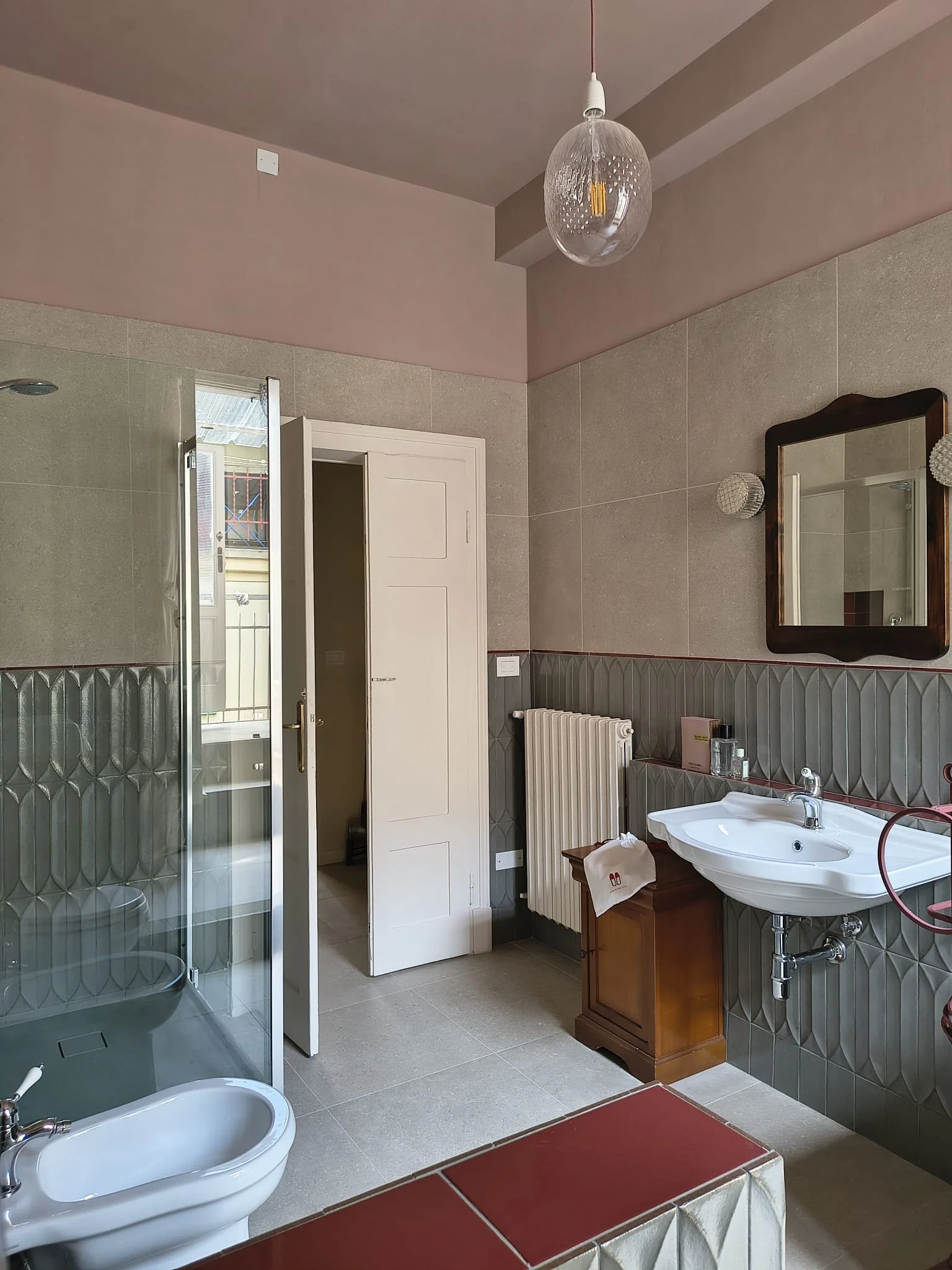 Una stanza da bagno con lavabo, grande specchio, radiatore e cabina doccia, pareti rivestite in piastrelle grigie e clinica, soffitto rosa antico con lampada appesa.