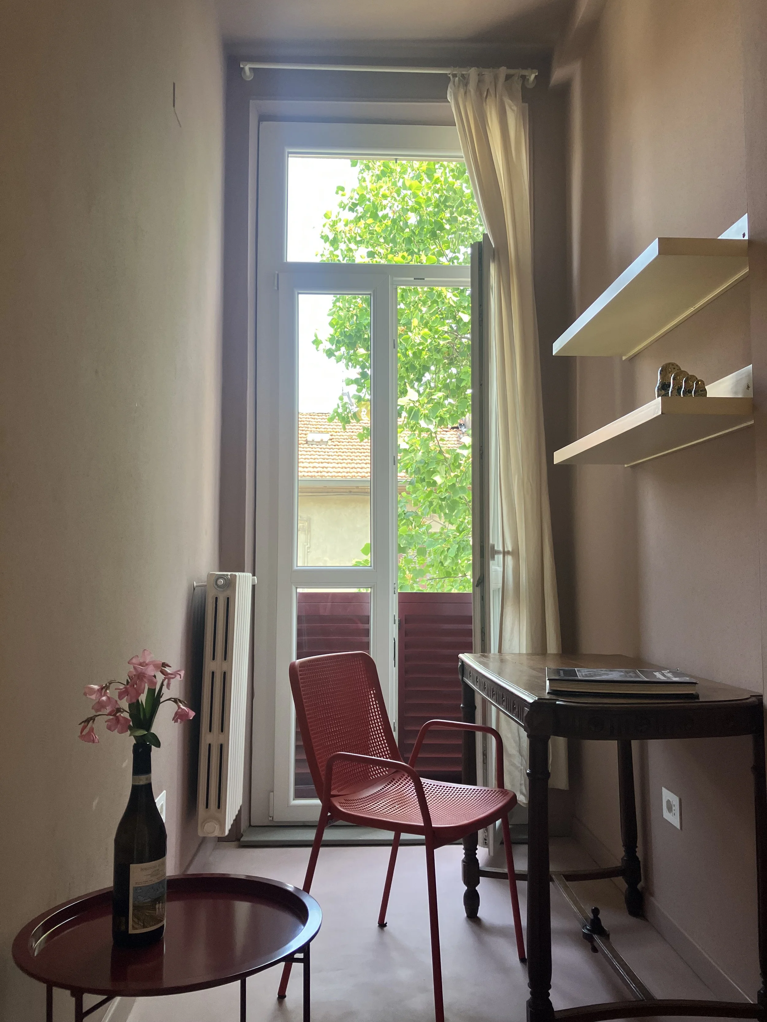 Interno di una stanza con finestra che mostra alberi e tetti di case. Sopra il tavolo ci sono due mensole vuote e sul tavolo ci sono alcuni libri. C'è una sedia rossa di plastica e un tavolino con una bottiglia di vino con fiori rosa.