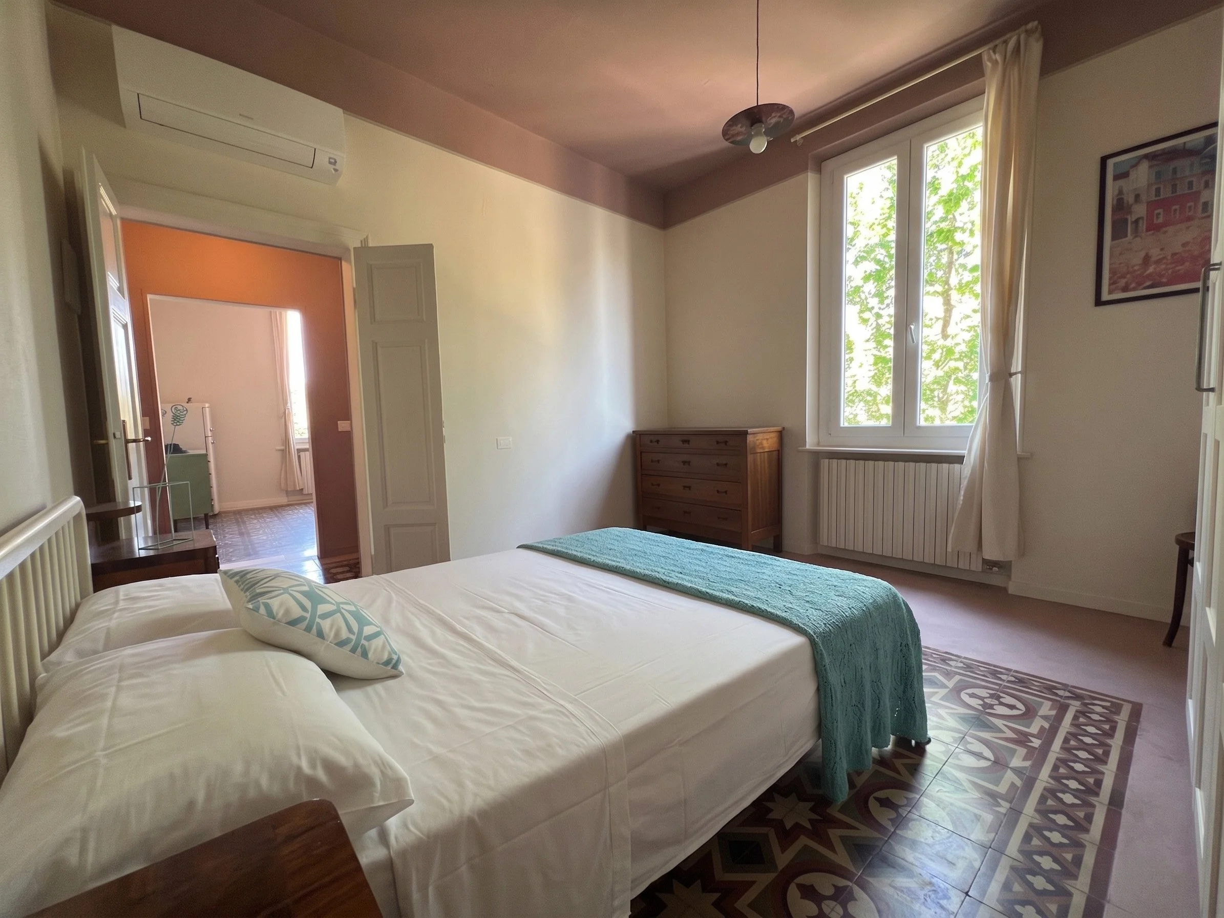 Camera da letto luminosa con un letto matrimoniale, armadio in legno e finestra con tende chiare, pavimento in piastrelle decorative.