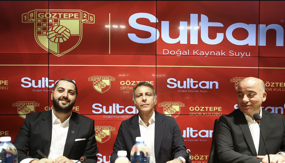 Three men in suits sitting at a table with water bottles in front of a red backdrop featuring logos of Göztepe Sports Club and Sultan natural spring water. The backdrop displays the Göztepe emblem and the words 'Sultan' and 'Doğal Kaynak Suyu' multiple times.
