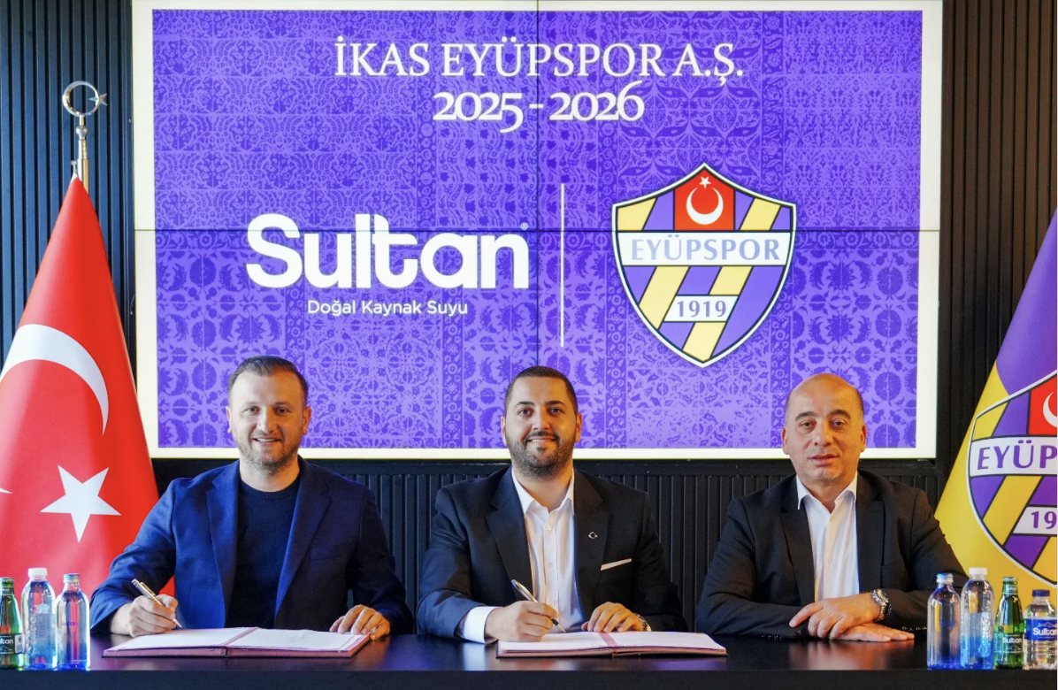 Three men in formal attire sitting at a table signing documents in front of a large screen that displays the logos of İkas Eyüpspor and Sultan Natural Spring Water. The Turkish flag and another flag with the Eyüpspor logo are visible on either side of the table.