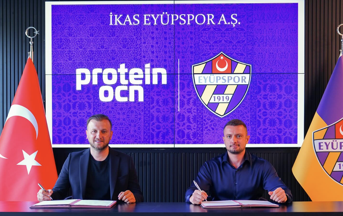 Two men sitting at a table with open documents, signing agreements, in front of a backdrop with the logos of Protein Ocn and Eyüpspor, a Turkish football club. The Turkish flag is on the left and Eyüpspor flag on the right.
