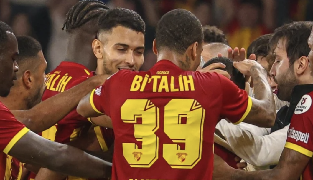 Soccer players in red jerseys celebrating on the field, with some pushing or touching each other's heads or shoulders.