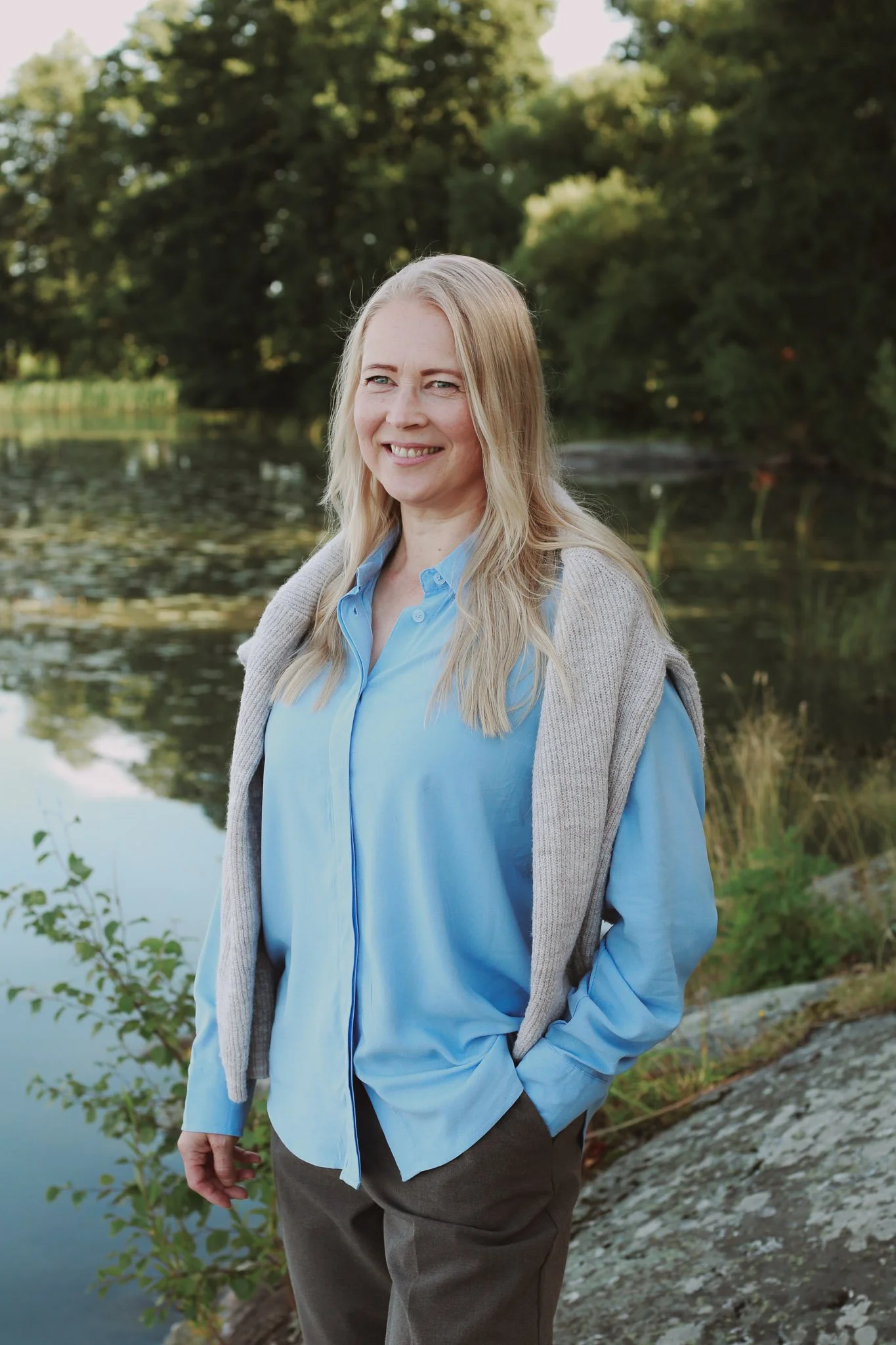 Mira Mustonen seisoo järven rannalla, taustalla puita ja vesistö, hänellä on vaaleat hiukset ja hän hymyilee.