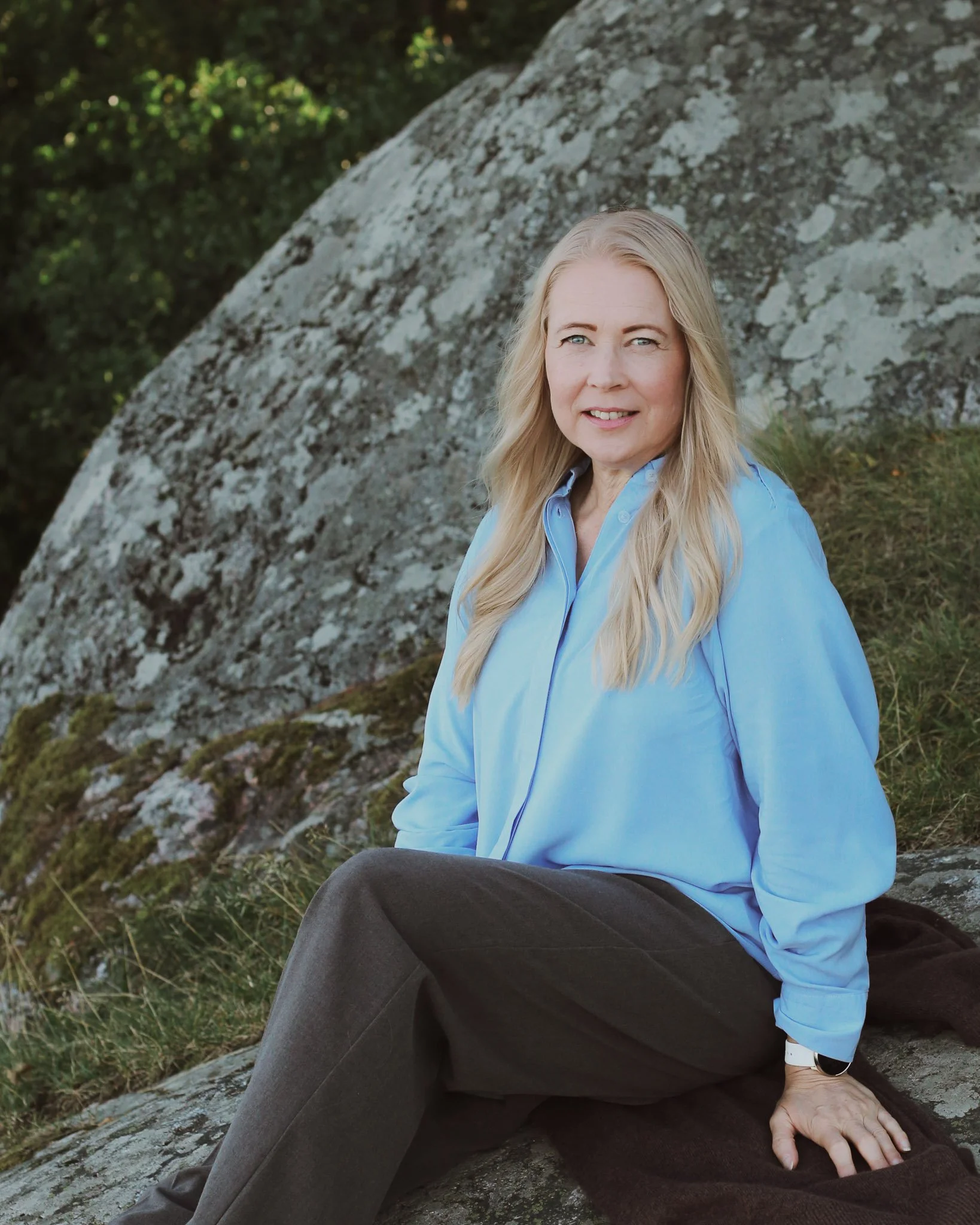 Mira Mustonen istuu rinteessä luonnon helmassa, taustalla puita ja kiviä, hänellä vaaleat hiukset ja sininen paita.