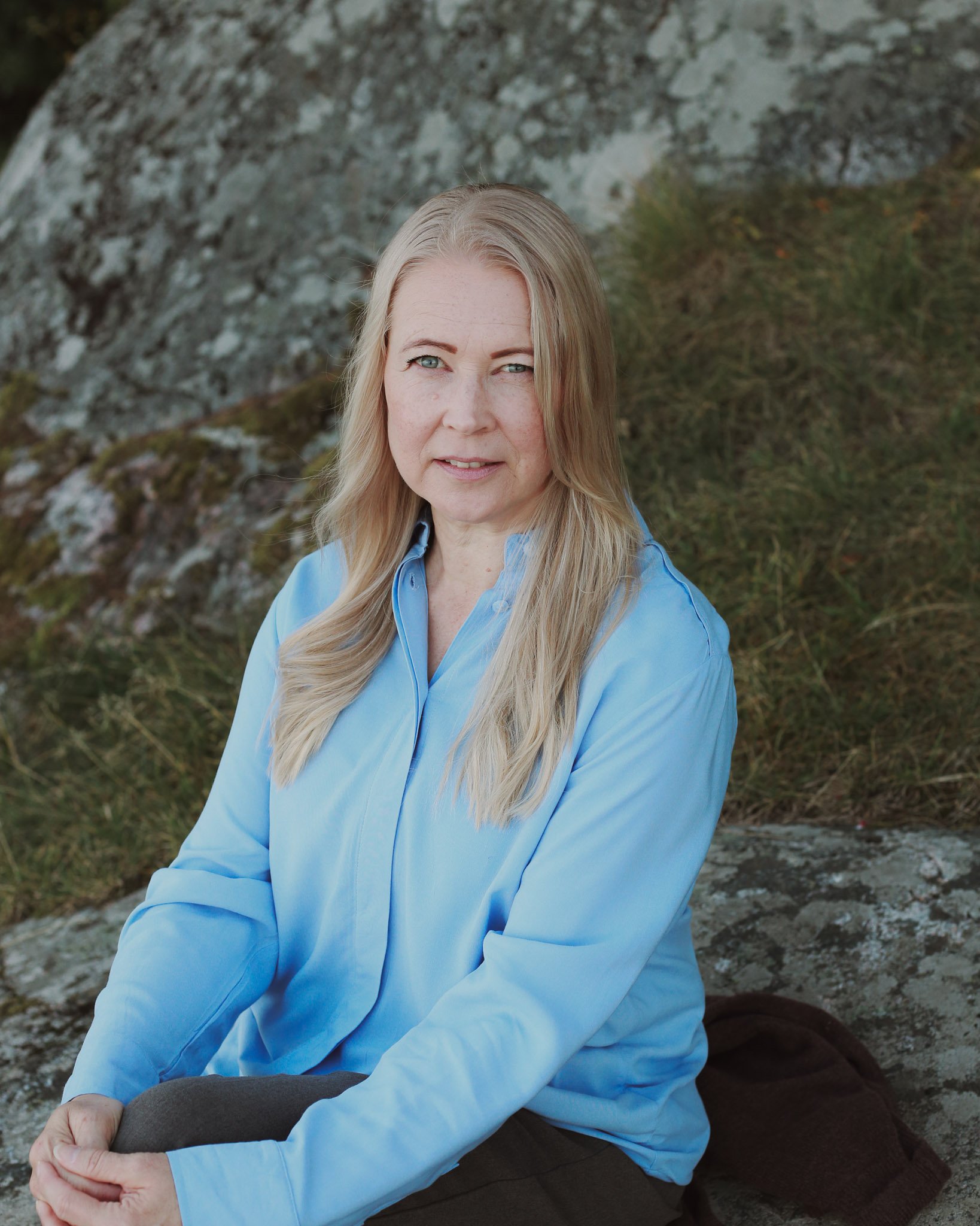 Kuvassa Mira Mustonen, jolla on vaaleat hiukset ja vaaleansiniset puserot, istuu ulkona kivisellä ja kasvillisuuden ympäröimällä alueella.