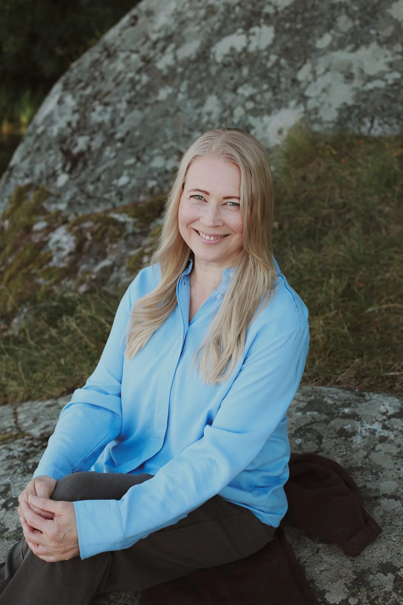 Mira Mustonen istuu kivellä ulkona luonnossa, taustalla suuri kivi ja vihreää kasvillisuutta. Hän on iloinen, hymyilee ja katsoo kameraan.