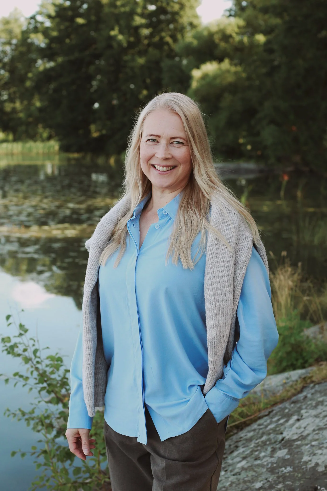 Mielen mietteet yrittäjä luonnonkauniissa ympäristössä, järven rannalla, vihreitä puita taustalla, hymyilee kameralle.