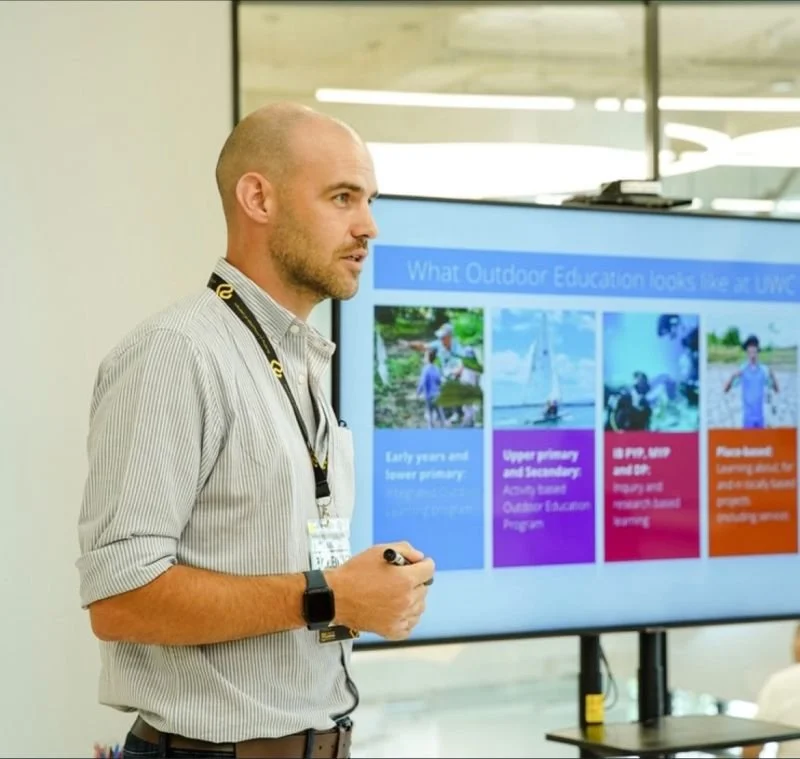 Man giving a presentation in front of a digital screen with slides on outdoor education.