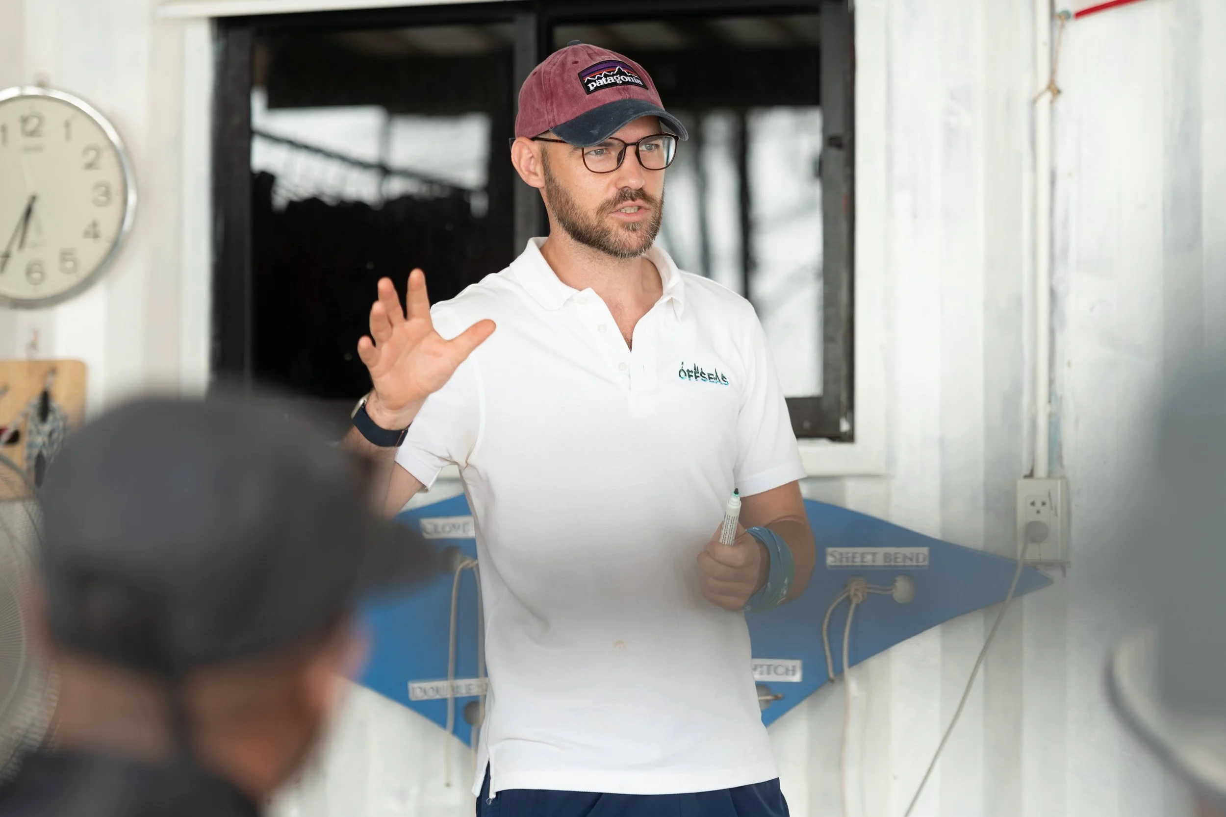 A man wearing glasses, a maroon cap, and a white polo shirt speaking and gesturing in a room with a clock and blue work station labeled "Sheet Bend" and "Mooring," holding a marker in his right hand.