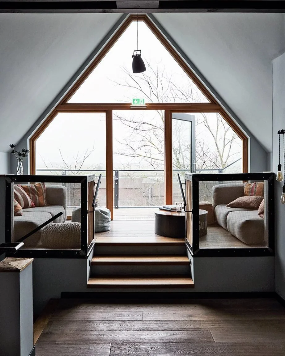 Ein gemütlicher Raum mit großen Fenster im Dachgeschoss, zwei Sofas mit Kissen, ein Couchtisch mit Büchern, und eine Hängelampe. Der Blick geht nach draußen auf eine Winterlandschaft mit Bäumen.