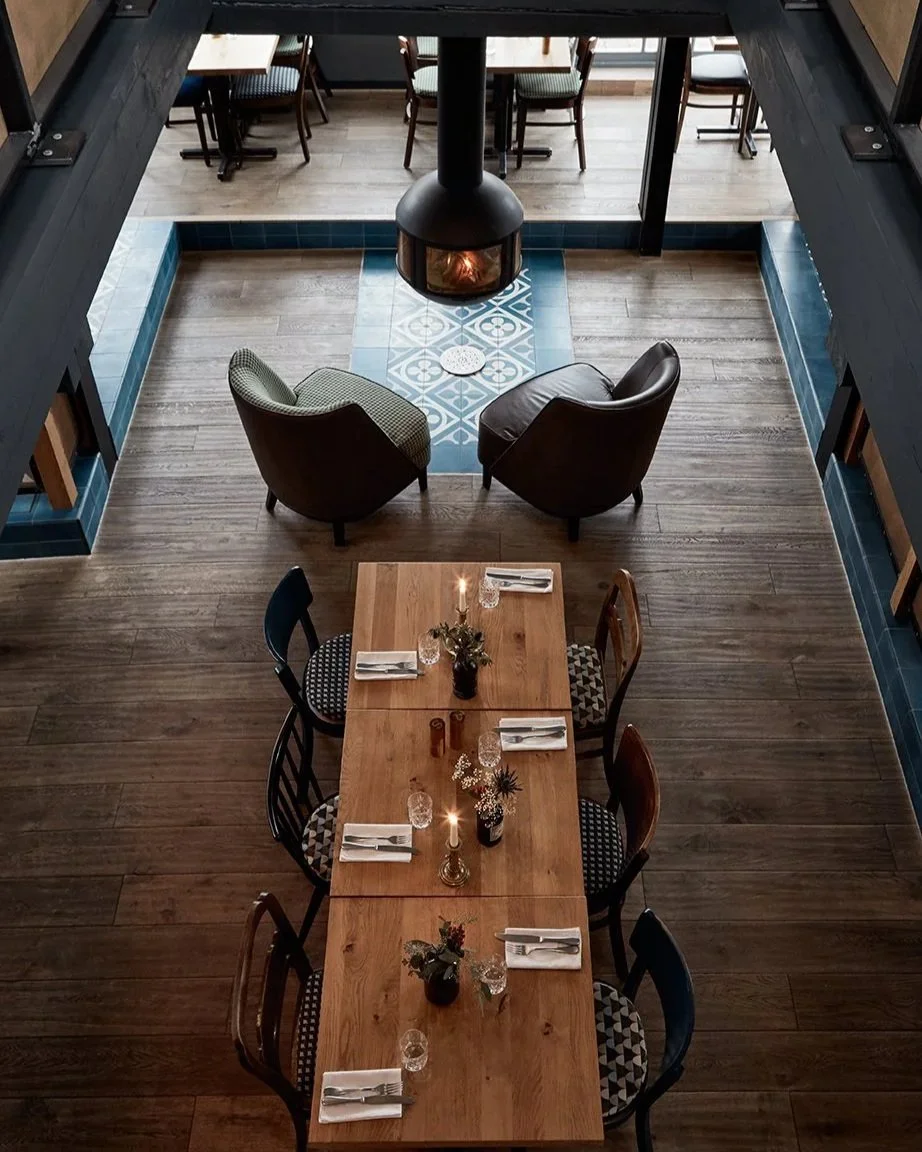 Gemütlich eingerichteter Restaurantbereich mit Holztisch, dekorierten Stühlen, Kerzen und Gläsern, Blick auf einen Kamin.