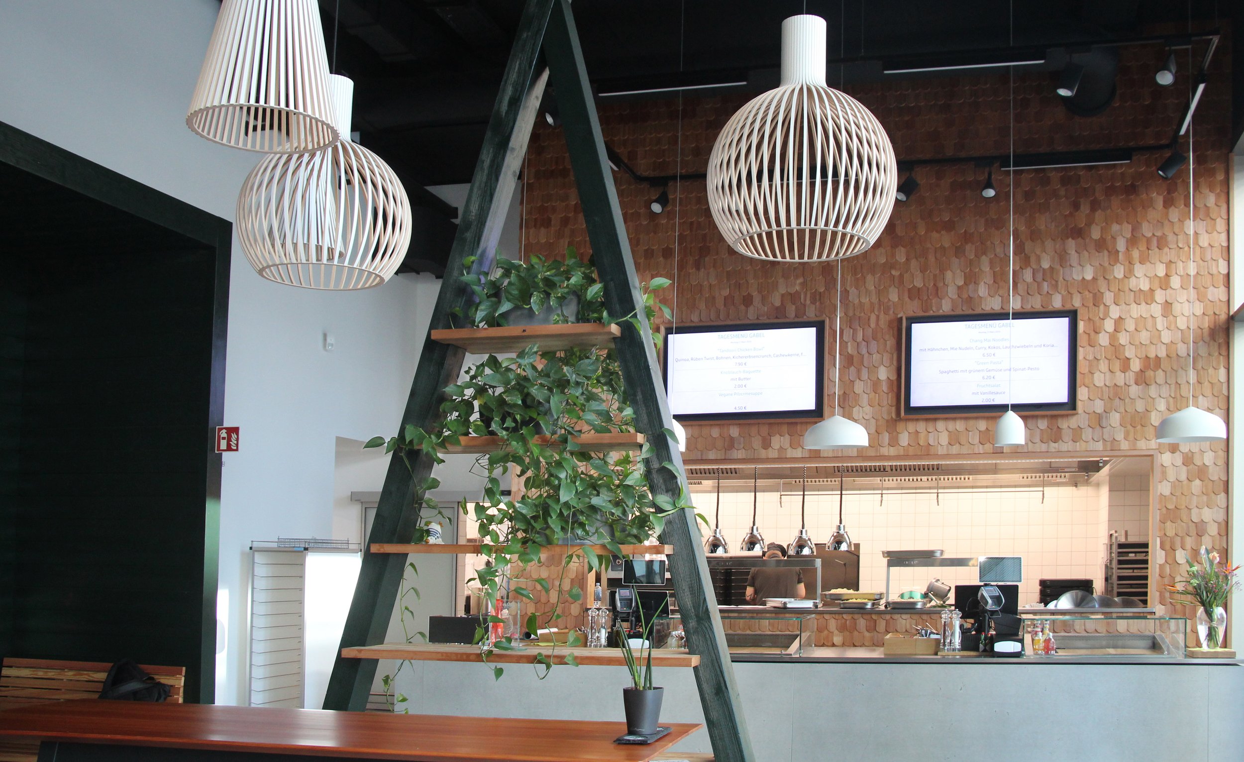 Innenansicht eines modernen Restaurants mit Hängeleuchten, grüner Pflanze auf einer Leiter, digitaler Speisekartenanzeige an der Wand, offener Küche im Hintergrund und einem Holz-Wartebereich.