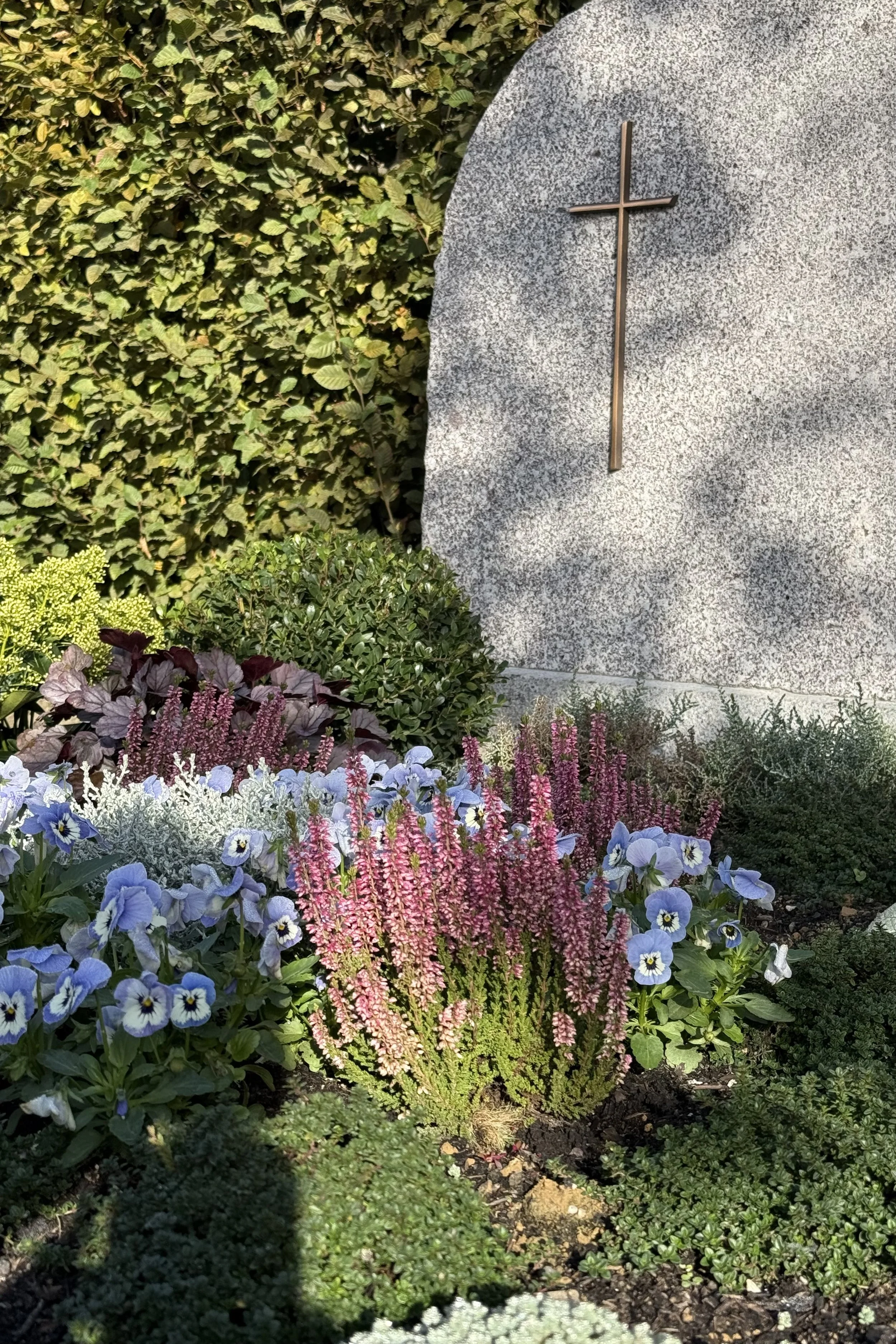 Blumen im Garten, darunter Vergissmeinnicht, Schleifenblumen, Fetthenne, Stauden, im Hintergrund eine Steinskulptur mit Kreuz