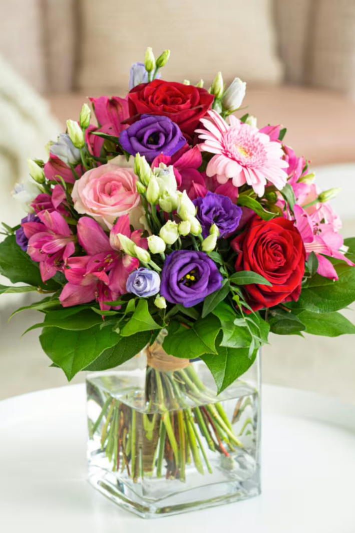 Bunte Blumenstrauß mit roten, pinken, lila und weißen Blumen in einer klaren Glasvase auf einem weißen Tisch.