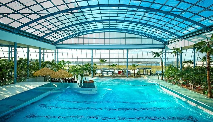 Innenansicht eines Schwimmbads in einer großen, glasüberdachten Halle mit Palmen und Sonnenliegen.