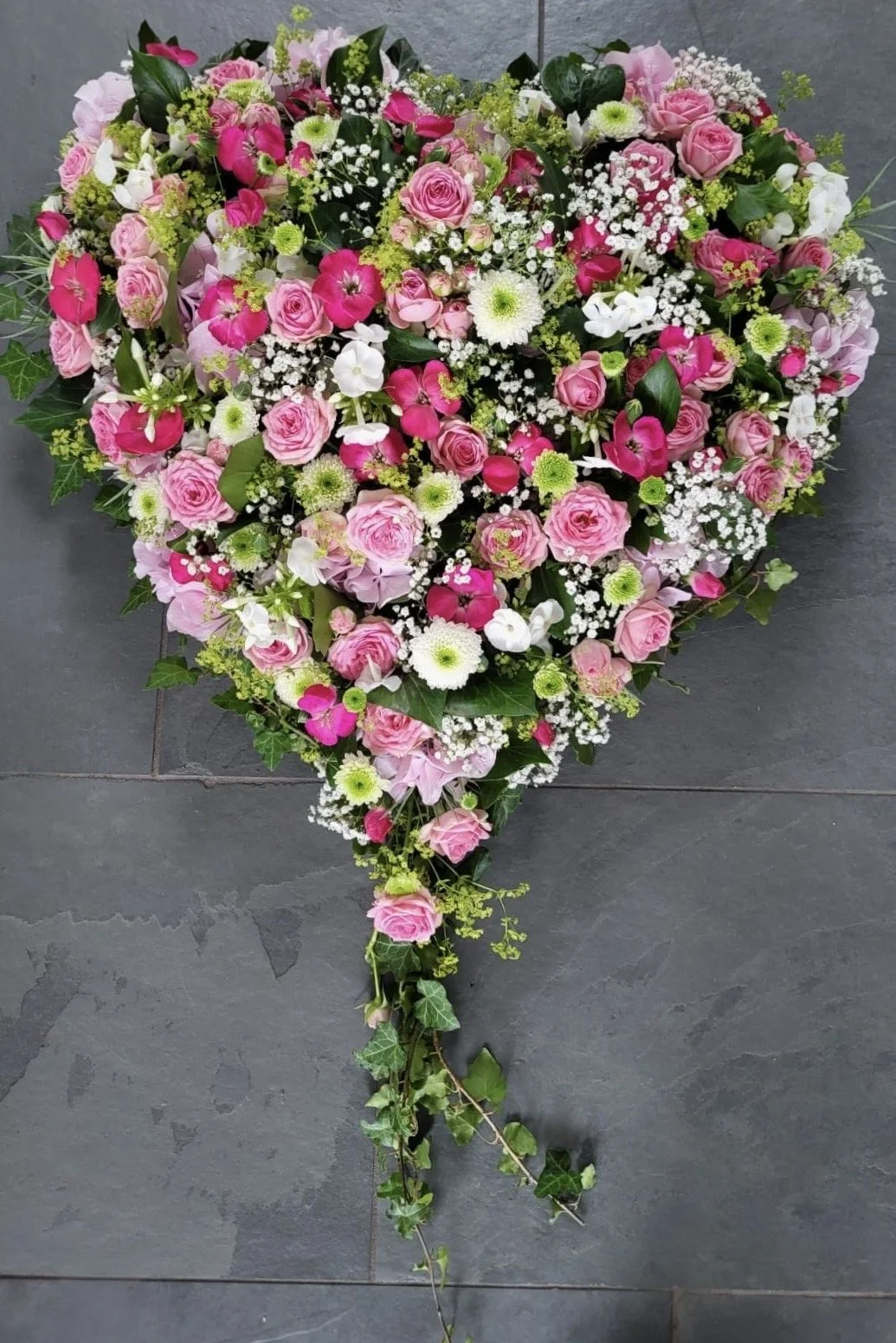 Ein herzförmiger Blumenstrauß aus rosa und weißen Blumen, einschließlich Rosen, Gänseblümchen und Veilchen, mit grünen Blättern auf einem grauen Boden.