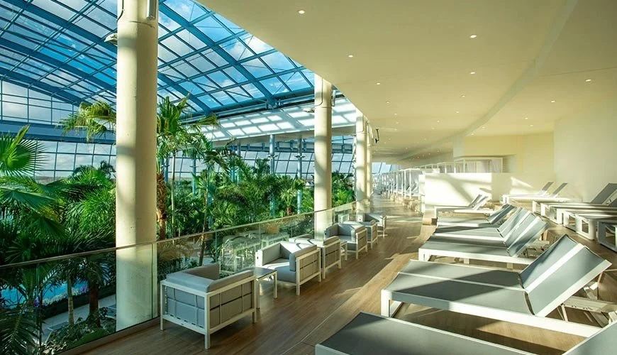 Innenraum eines modernen Wellnessbereichs mit Sonnenliegen, großen Fenstern, die einen Wintergarten mit vielen Pflanzen zeigen, und einer hellen, offenen Architektur.