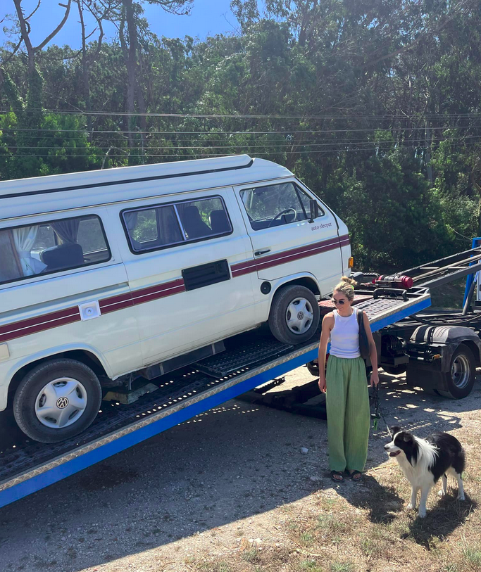 A woman with sunglasses and casual clothing stands next to a black and white dog, holding its leash, beside a flatbed truck containing an older Volkswagen camper van, with trees and power lines in the background.