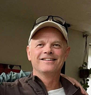 A smiling man wearing a baseball cap and sunglasses on top, with a relaxed background.