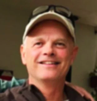 A smiling man wearing a baseball cap with sunglasses resting on top, indoors.
